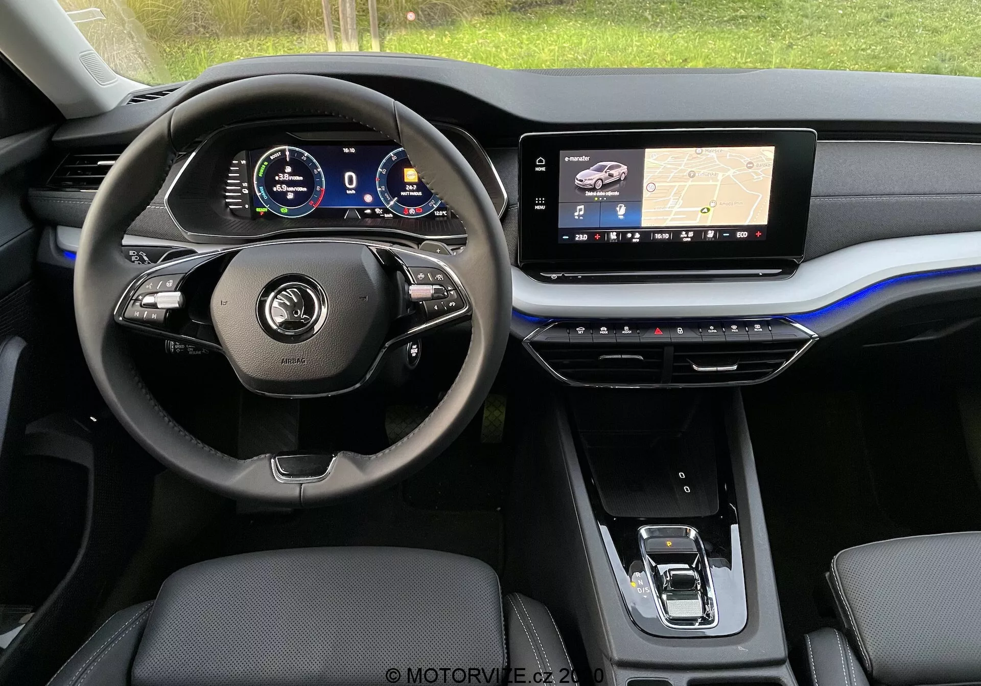 Egy modern Skoda Octavia Limousine 2020 belső nézete a hátsó ülésből, amely az első műszerfalat és a vezetőülés területét mutatja. A kormánykerék többgombos vezérlőkkel és bőrborítással rendelkezik, mögötte egy digitális műszercsoport található, amelyen különböző járműmérők láthatók. A középső műszerfal nagy infotainment érintőképernyőt tartalmaz, amely egy navigációs térképet jelenít meg GPS technológiával, míg az alatta lévő középső konzol érintésérzékeny gombokat tartalmaz a klímavezérléshez. Láthatók a bőrborítású első ülések és egy elektronikus sebességváltó. Az esztétika elegáns, sötét tónusokkal, fémes kiemelésekkel és egy merész kék sávval a műszerfalon.