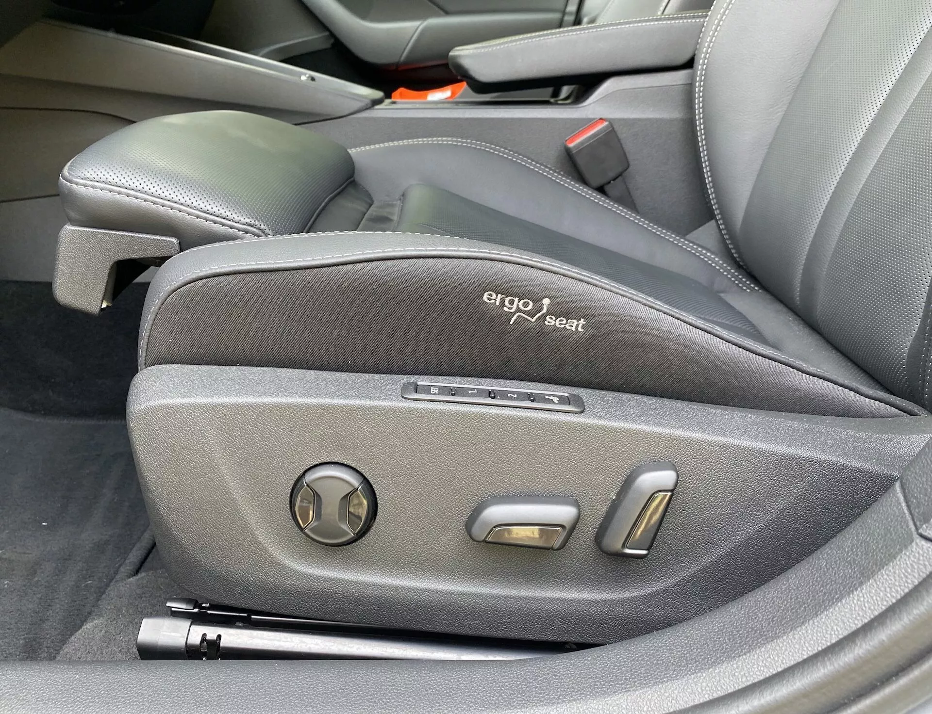 A 2020-as Skoda Octavia vezetői ülés oldalirányú vezérlőinek közelről készült képe, amely ergonómikus kialakítást mutat sötét szövetkárpitozással, látható 'ergo seat' jelzéssel és elektronikus állító vezérlőkkel, beleértve egy kör alakú dőlésszögállítót és két fémes helyzetbeállító gombot, kiemelve a jármű prémium és technológiailag fejlett belsőterét.