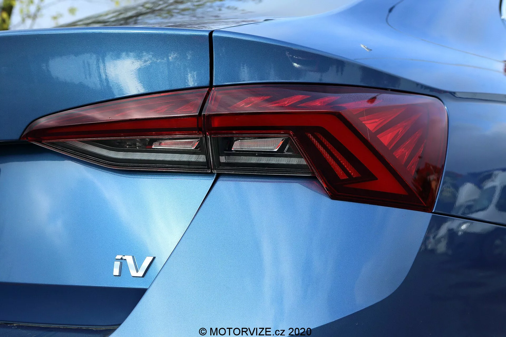 2020-as Skoda Octavia Limousine hátsó részének közeli részlete, amely elsősorban a bal oldali hátsó lámpa szerelvényre összpontosít, háromnegyedes szögből nézve. A modern hátsó lámpa dizájn éles szögekkel és jellegzetes LED világító elemekkel rendelkezik, amelyek zökkenőmentesen integrálódnak az autó karosszéria vonalaival. A fényes, fémes kék csomagtérfedélen látható 'iV' jelvény hibrid vagy elektromos járművet sugall.