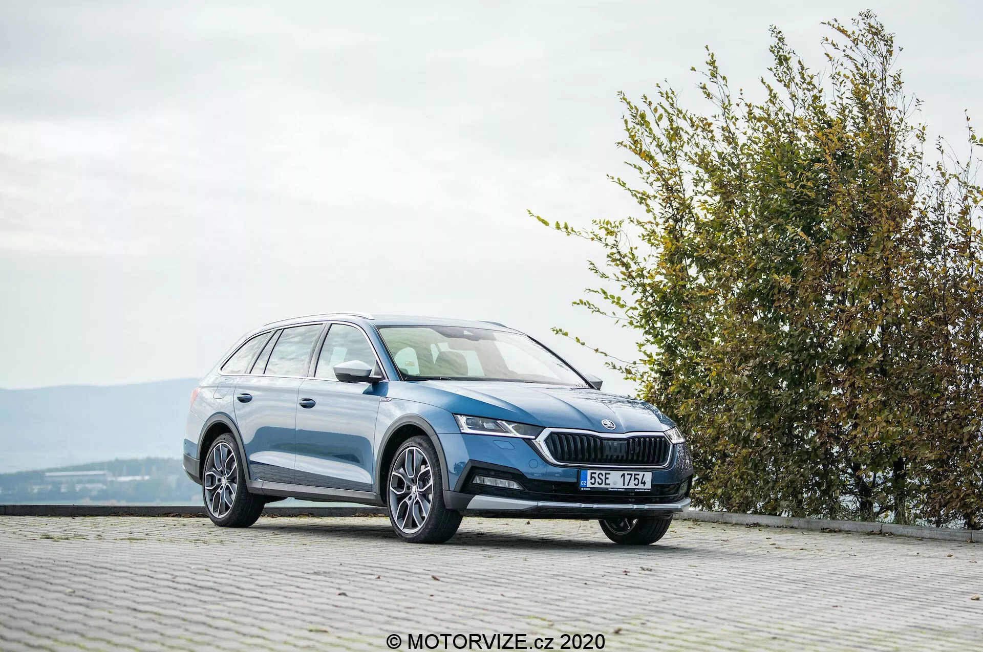 2020-as Skoda Octavia Scout teljes méretű kombi autó, háromnegyed elölnézetből fényképezve burkolt, macskaköves területen, egy emelt helyszínen. A kép bemutatja az elülső hűtőrácsot feltűnő emblémával, elegáns, szögletes fényszórókat, integrált ködlámpákat, sima oldalsó profilt könnyűfém kerekekkel, tetősíneket és fényes festékbevonatot. A környezet része egy sövény vagy bokor a jobb oldalon, amely park vagy zöldterület közelségét jelzi, kedvező időjárási körülmények között.