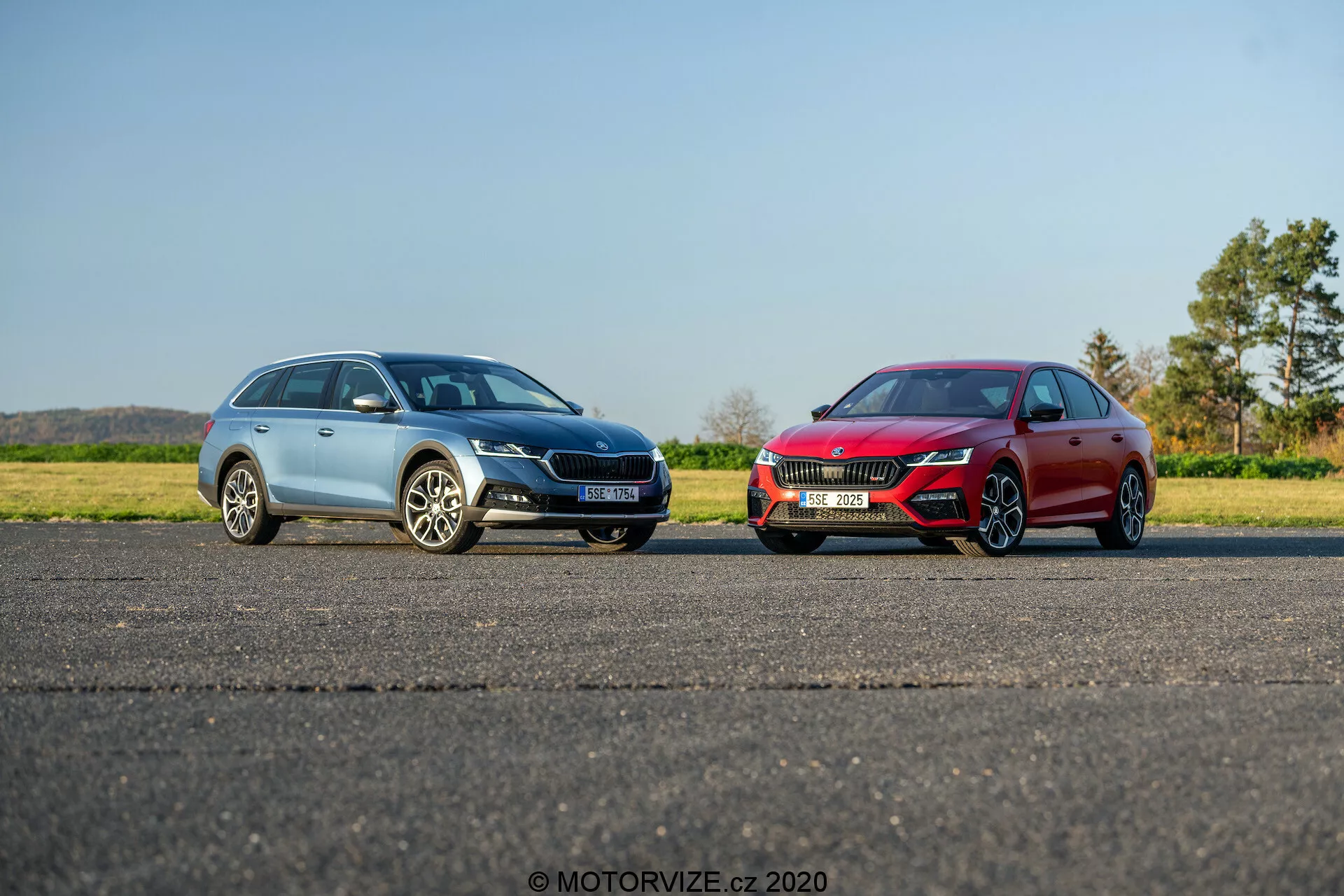 Két autó parkol egymás mellett egy sík aszfaltos felületen, tiszta kék ég alatt, vidéki vagy félvidéki környezetben. A bal oldali kék SUV többküllős alufelniket, elegáns LED fényszórókat, jellegzetes hűtőrácsot és látható tetősín található. A jobb oldali piros sportos szedán alacsonyabb profilú, nagyobb sportfelniket, agresszív LED fényszórókat, széles hűtőrácsot és elegáns, aerodinamikus első lökhárítót tartalmaz. Mindkét jármű valószínűleg modern technológiai funkciókkal van felszerelve. A fényképen nincs mozgás elmosódás, az árnyékok arra utalnak, hogy a fotó reggel vagy késő délután készült. A „MOTORVIZE.cz 2020“ vízjel jelzi a képet forrását és évét.
