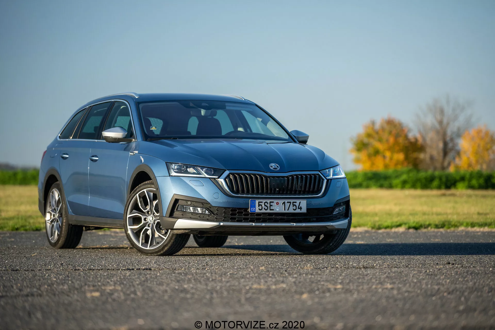 Egy modern, kék kombi háromnegyedes elülső nézetből készült képe mutatja be elegáns, korszerű kialakítását. A jellemzők között szerepelnek a szögletes fényszórók LED-es nappali menetfénnyel, a króm bevonatú, részletes vízszintes hűtőrács, a többrudazatos könnyűfém felnik, a beépített irányjelzővel ellátott oldalsó tükrök, az alacsony profilú gumiabroncsok, a tetősín és a sötétített hátsó ablakok. Az autó egy burkolt úton van elhelyezve, háttérben őszi lombkoronával, alacsony szögből fényképezve, hogy kiemelje a dizájnvonalakat és a hasmagasságot. A természetes nappali fény kiemeli a jármű fényezésének kidolgozottságát és a dizájn részleteit.