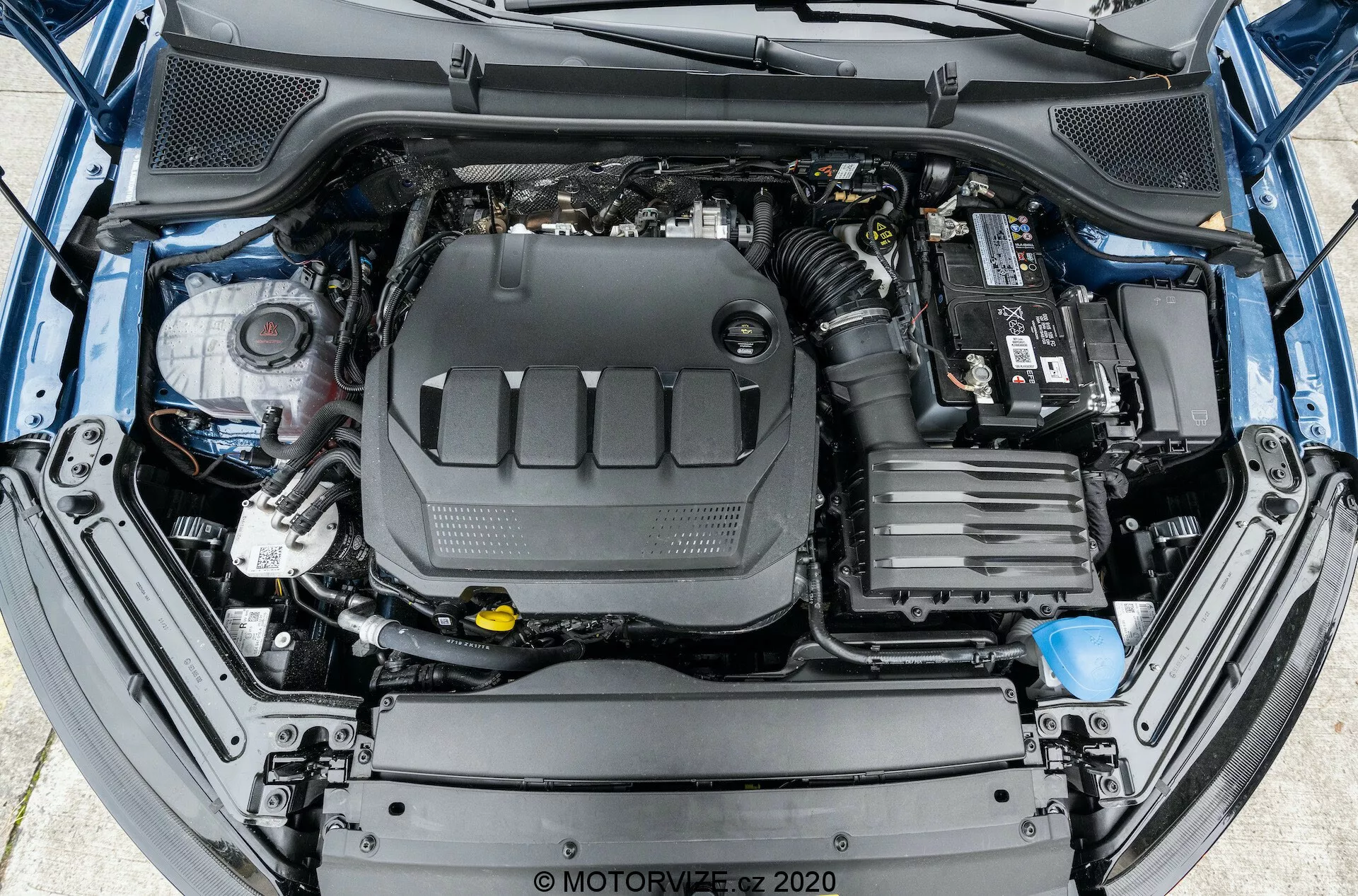 Madaras szemszögből készült kép egy nyitott autómotor éveleiről, amely a motor és környező alkatrészeinek átfogó elrendezését mutatja. A motorblokk központi helyzetben van, műanyag motorborítással. Jobbra, az autó akkumulátora látható a terminálkapcsolatokkal a tetején. Előre, a jármű légbeömlő rendszerének alkatrészei láthatók. A bal oldalon különféle folyadéktartályok vannak, például a fékfolyadék tartálya sárga fedéllel. A motor room térben különféle csövek és vezetékkötegek is láthatók. Az első lökhárító és rácsszerkezet részben látható a kép alsó szélén.