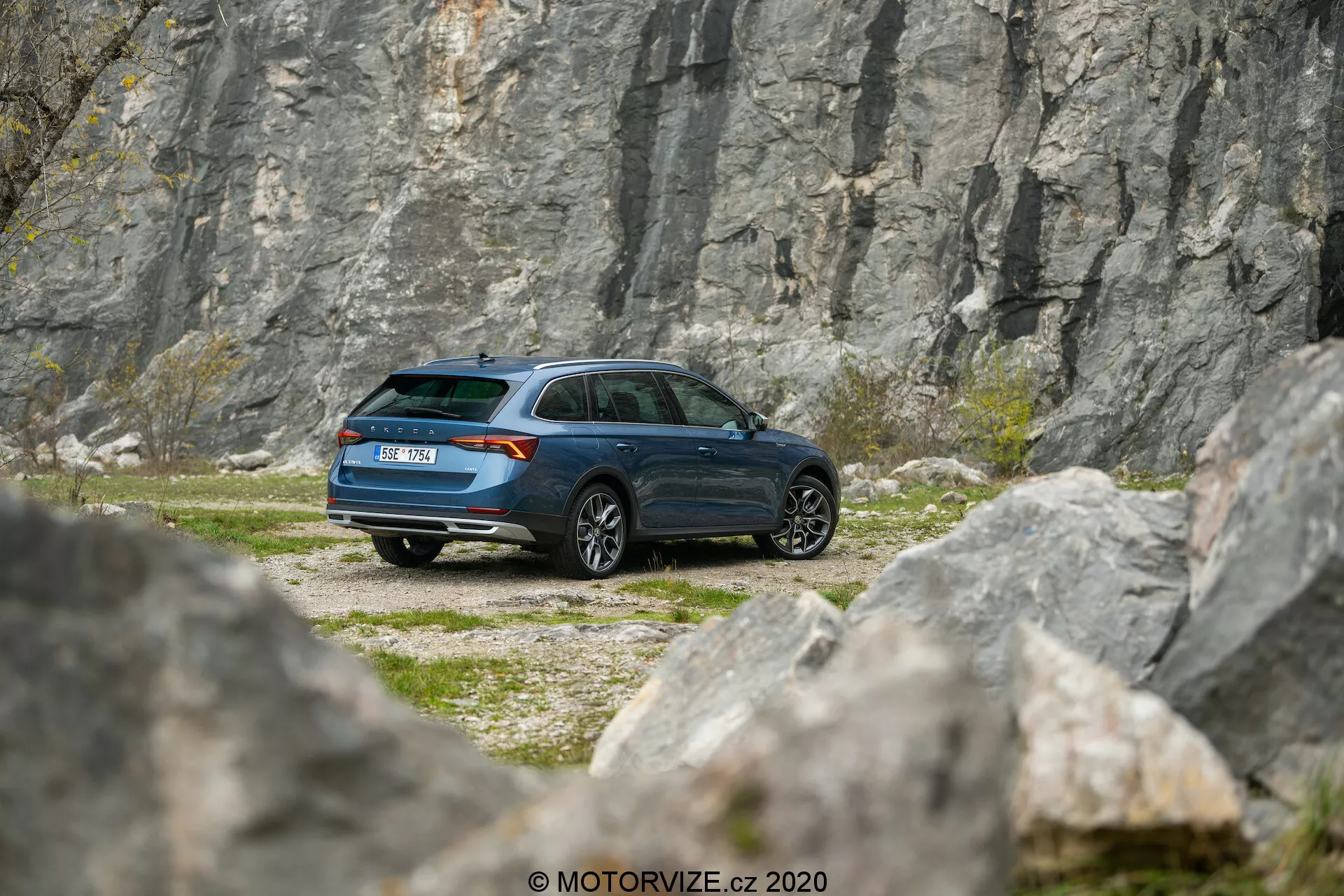 A képen egy 2020-as Skoda Octavia Scout látható, amely sziklás terepen parkol, háttérben egy kőfejtővel vagy sziklafallal, enyhe teleobjektív perspektívából megörökítve. A háromnegyedes hátsó szög részletes áttekintést nyújt a jármű oldalsó profiljáról és hátuljáról, bemutatva a modern kombi kialakítást, a ferde hátsó tetővonalat és a sötétkék festést. Látható jellemzők közé tartozik a hátsó lámpatest, az ablakok formája, a hátsó ablaktörlő, a kerekek kialakítása és az oldalsó tükrök. A környezet a kaland és felfedezés témáját idézi, kiemelve az autó képességét távoli és nehéz terepek elérésére.