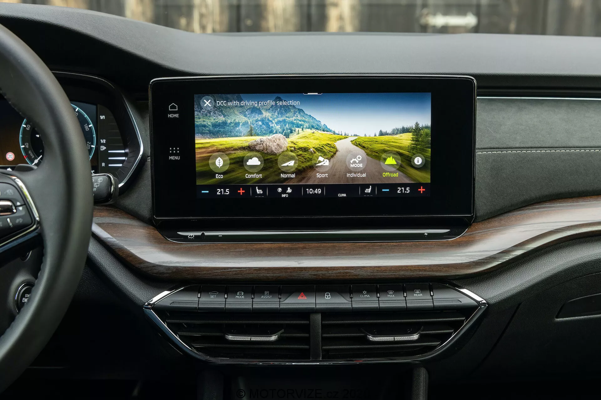 A Skoda Octavia 2020 (Octavia Scout) belső nézete bemutatja a modern szórakoztató rendszert és a műszerfalat, a hátsó utastér perspektívájából fényképezve. A műszerfalon található széles, vízszintesen tájolt érintőképernyő színes grafikákat, klímavezérlő beállításokat, időt és külső hőmérsékletet mutat. Alatta rendezetten elhelyezett klímavezérlő gombok és kapcsolók láthatók. A luxus műszerfal puha tapintású anyagból és fa furnérból készült, diszkréten beépített szellőzőnyílásokkal.