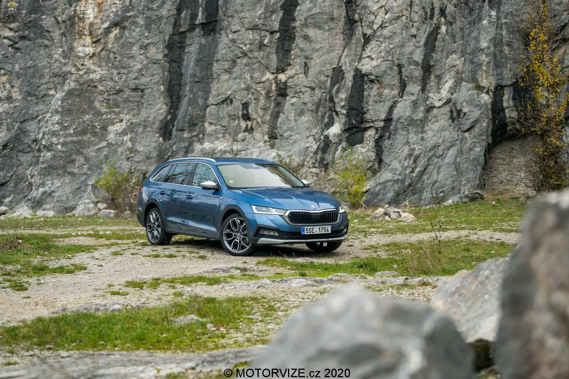 Oldal-elölről nézve egy kék Skoda Octavia Scout 2020 kombi kavicsos terepen, sziklás háttérrel, bemutatva a modern LED fényszórókat, a kiemelkedő hűtőrácsot, az alumínium kerekeket, az oldalsó tükröket, ablakdíszlécet, tetősíneket és a tiszta metálos felületet, amely fejlett funkciókat, például világítástechnológiát, gumiabroncsnyomás-figyelést és esetleges biztonsági rendszereket sugall.