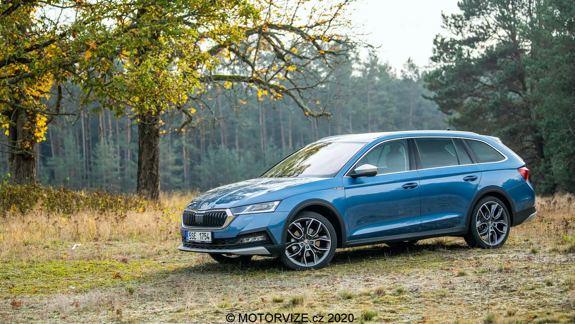 Az oldalnézeti képen egy kék 2020-as Skoda Octavia Scout állomáskocsi látható földúton, a háttérben fákkal és fűvel, bemutatva a terepjárás képességét. Az autó fémes festéssel, többrudas könnyűfém keréktárcsákkal, LED-es fényszórókkal, jellegzetes első ráccsal, tetősínekkel, karosszéria színű külső tükrökkel és ajtókilincsekkel, valamint napfénytetővel rendelkezik. A tervezési jellemzők között szerepel a karaktervonal, amely az első kerékívtől a hátsó lámpákig tart, és az ölelő hátsó lámpák is.