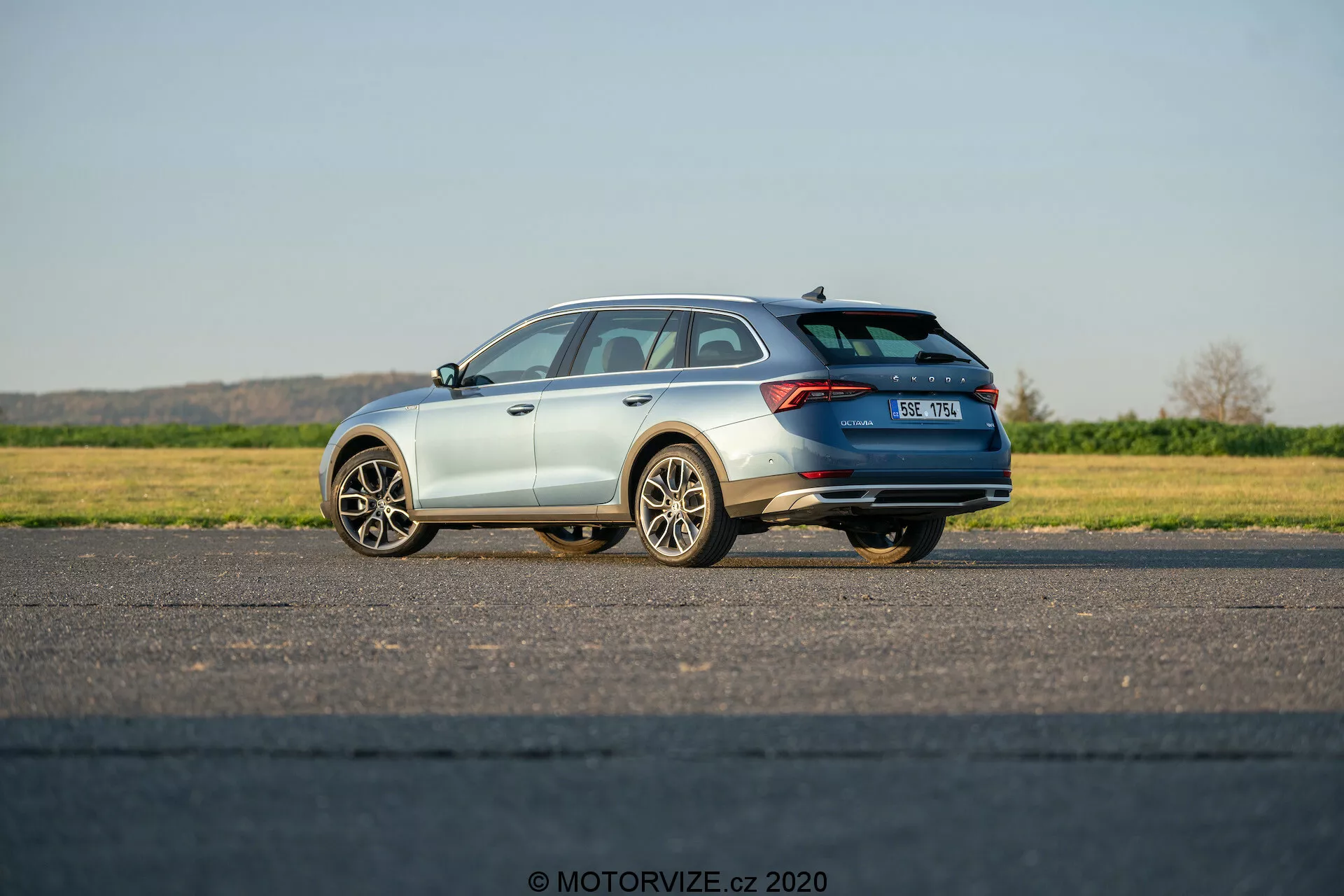 A képen egy 2020-as Škoda Octavia Scout látható, egy modern kombi, háromnegyedes hátsó nézetből fényképezve. Kiemeli az autó dizájnelemeit, beleértve a vízszintesen elhelyezkedő hátsó lámpákat, amelyek a csomagtér közepéig nyúlnak, a tetőspoiler, a tetősínek és a többküllős alufelnik. Az autó egy sík, burkolt felületen áll, a vezető oldala közelebb helyezkedik el a kamera felé. Az autó fémes festékréteggel van borítva, és látható a kipufogórendszer egy része. A rendszámtábla kissé elmosódott, és a Škoda jelvény látható. A természetes nappali fényben, tiszta égbolt alatt készített fotó egy álló járművet mutat, amely potenciálisan rendelkezhet rejtett technológiai funkciókkal, mint például parkolássegítő rendszer, hátsó kamerák vagy ütközéselkerülő rendszerek.