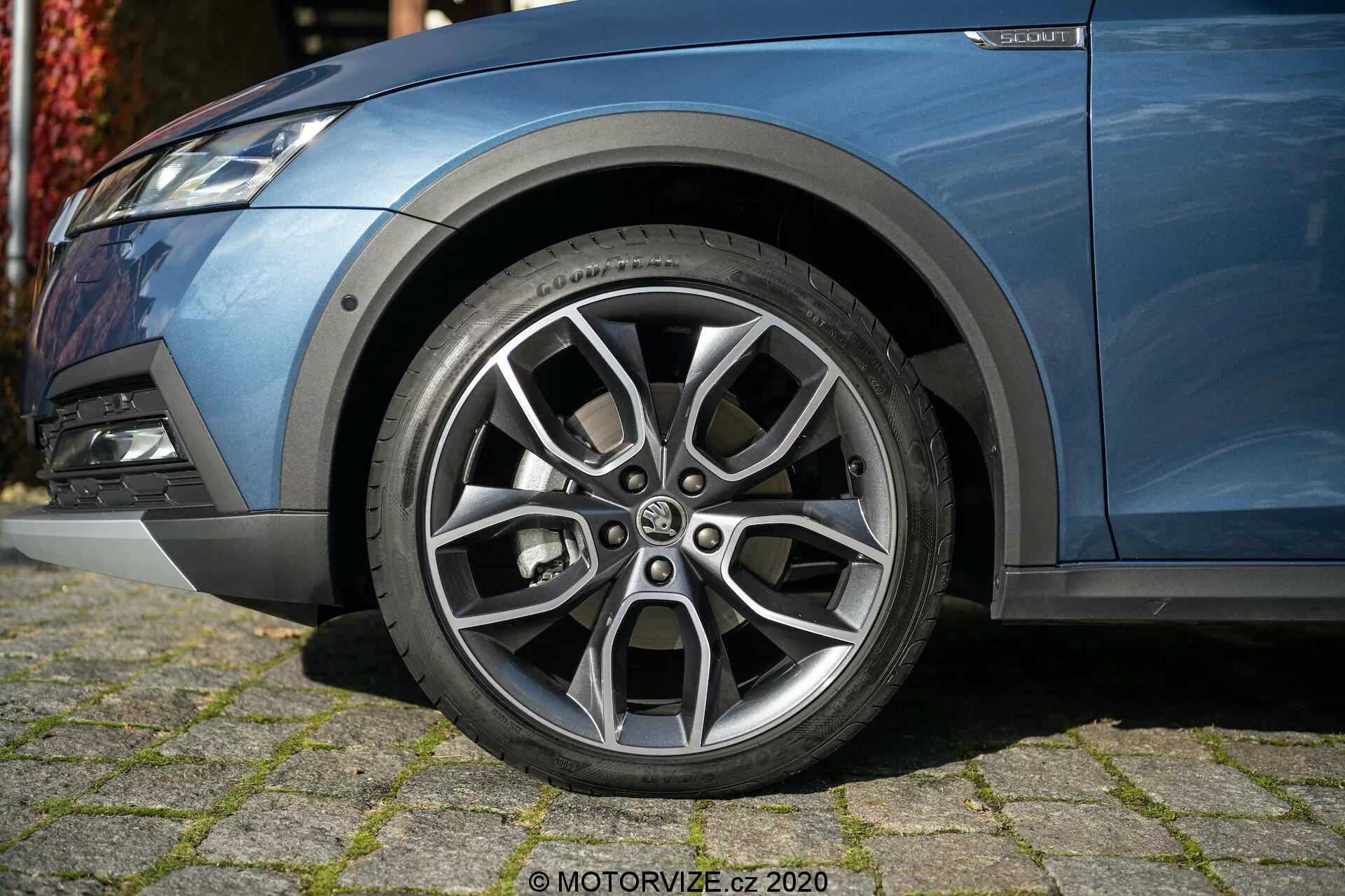 Közeli kép egy Škoda Octavia 2020 (Octavia-Scout) bal első oldaláról, kiemelve a gondosan megtervezett, többszálú kialakítású, ezüst színű és kontrasztos felületű könnyűfém kereket, valamint a látható futófelület mintázatot. Az alacsony szögből készült fotó a kocsi kék, metálszínű karosszériájának egy részét, a ködlámpás első lökhárítót és a felfüggesztés elemeit is tartalmazza, bemutatva a modern, sportos megjelenést.