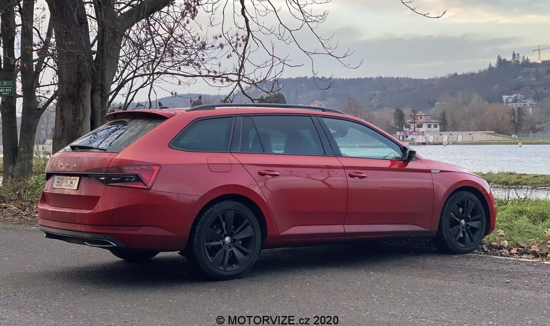 Egy piros Skoda Superb 2019 kombi parkol a szabadban, háromnegyedik hátsó szögből nézve, amely a kocsi jobb oldalát mutatja. A kulcsfontosságú tervezési elemek közé tartoznak a nagy, sötét színű könnyűfém kerekek, a lejtős hátsó és oldalsó ablakok, az átfogó hátsó lámpák, a zárt hátsó ajtó jelvénnyel, a diffúzoros és dupla kipufogós hátsó lökhárító, a tetősínek és a színre festett külső oldalsó tükör. A környezet zöld növényzetet és egy vízfelületet mutat a háttérben, ami egy nyugodt elővárosi vagy félvidéki környezetet sugall.