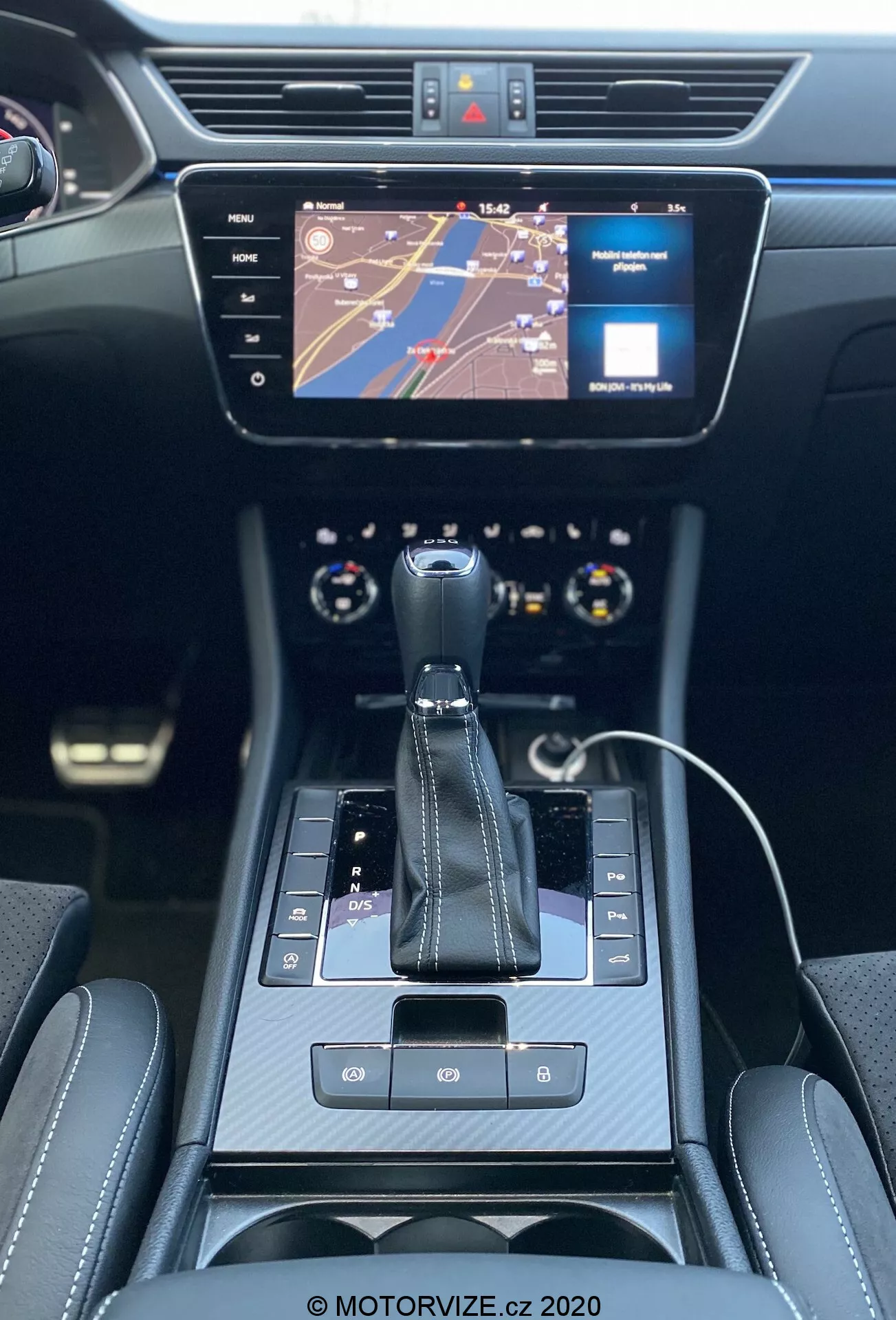 A 2019-es Skoda Superb Limousine belső nézete, középpontban a középkonzollal és az műszerfallal, hátsó ülés perspektívából. Automata sebességváltó kar 'P' parkoló jelöléssel, elektronikus rögzítőfék gomb, érintőgombok és érintőpad interface látható. Egy infotainment érintőképernyő a konzol felett térképet és médiajelzőt mutat. Kétzónás klímavezérlők a képernyő alatt, szellőzőrostélyokkal körülvéve. Modern, elegáns fekete belső tér krómozott kiegészítőkkel és látványos varrással ellátott luxus ülésekkel. Látható a multifunkciós kormány felső széle, valamint a műszerfal része analóg tárcsákkal. Okostelefon töltőkábellel utalva a csatlakoztathatósági funkciókra.