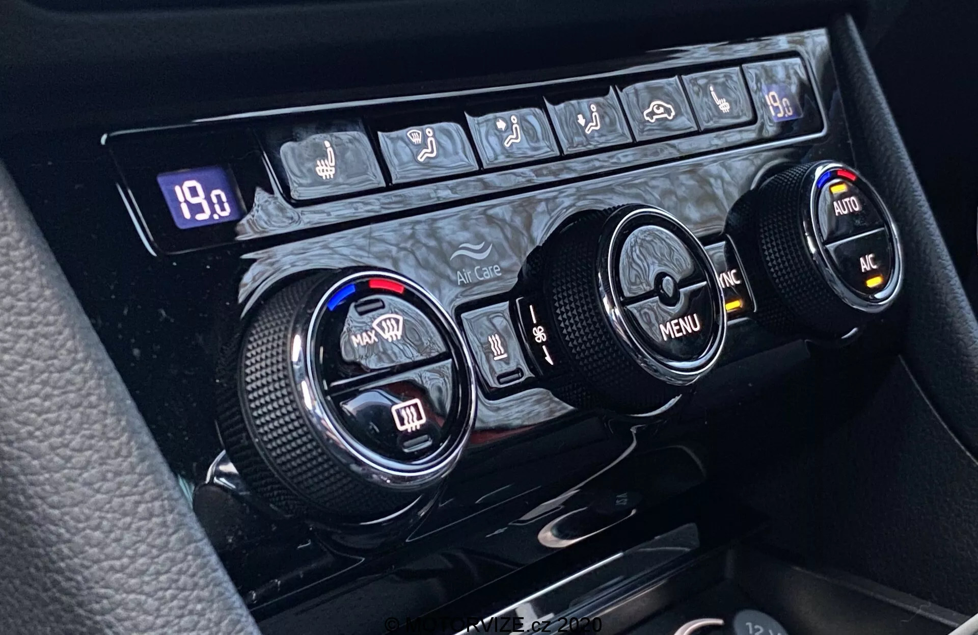 Közelkép a Škoda Superb 2019 klímaszabályozó rendszeréről, amely a középkonzolon helyezkedik el, három kör alakú tárcsával, amelyek a hőmérséklet-szabályozás, a menü navigáció, valamint további funkciókhoz használhatók. A bal oldali tárcsa a vezető, a jobb oldali pedig az utas számára állítja a hőmérsékletet, míg a középső tárcsa a menün való navigációt biztosítja irányított nyilakkal. A textúrált szélek ergonomikus használatot biztosítanak. A tárcsák felett egy sor gomb található, amelyek magukban foglalják a hátsó ablak páramentesítését, az első ablak páramentesítését, a levegő újrakeringetését, az automatikus klímaszabályozást, a légkondicionáló bekapcsolását és esetleg egy vészhelyzeti funkció gombot. Egy digitális kijelző mutatja a '19.0' értéket, ami a beállított hőmérsékletet vagy a rendszer működésével kapcsolatos egyéb értéket jelezheti.