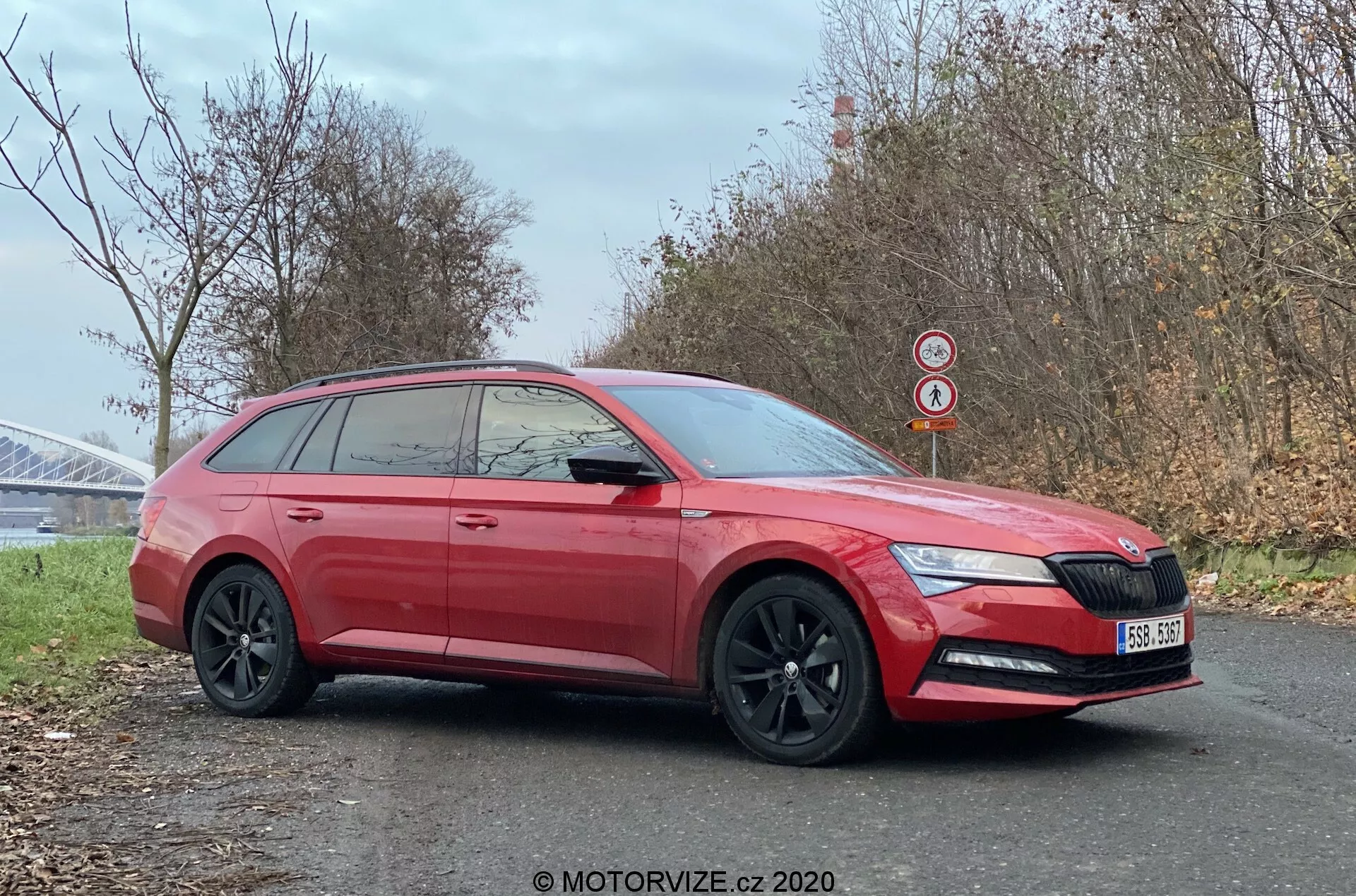 Egy piros 2019-es Skoda Superb kombi oldalsó profilja az út szélén parkolva, amely bemutatja modern kialakítását éles első fényszórókkal, beleértve a LED nappali menetfényeket, jellegzetes elülső hűtőrácsot logóval, fekete könnyűfém kerekeket, privát szférát biztosító sötétített ablakokat és a kombi karosszériára jellemző, meghosszabbított tetővonalat. A fotó úgy van keretezve, hogy kiemelje mind az oldalát, mind a kissé rövidített elejét, rögzítve olyan részleteket, mint az oldalsó tükrökbe integrált indexlámpák, a karosszéria színével megegyező ajtókilincsek, az alsó karosszéria burkolata és a hátsó tetőszpojler. A hideg évszakban, egy lehetséges híd szerkezete közelében készült tiszta kép láthatóvá teszi az autó jellemzőit és a környező környezetet.