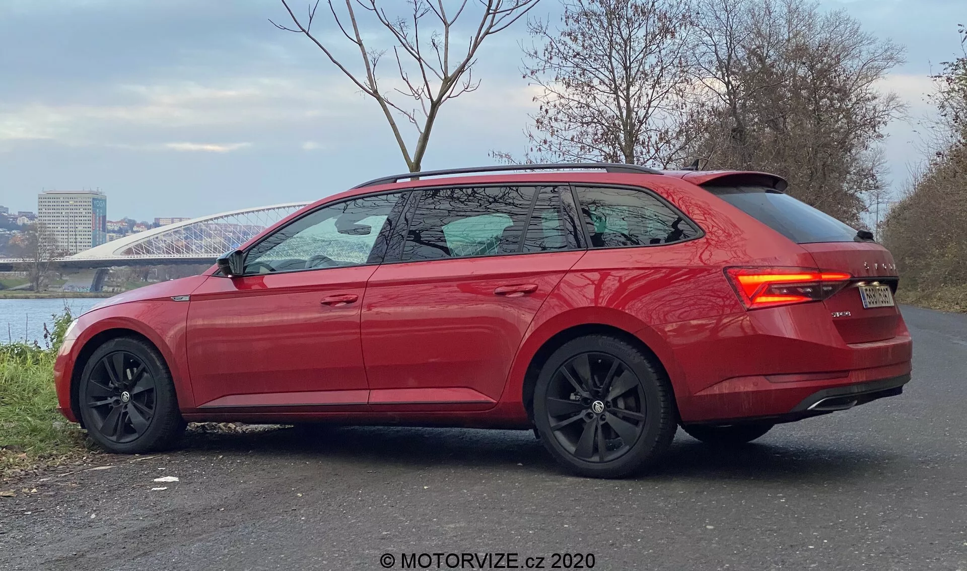 A kép egy modern, élénkpiros Skoda Superb 2019 kombit mutat háromnegyed hátulról nézve. Az autó egy kövezett területen parkol, háttérben folyóval és híddal, bemutatva a karcsú vonalakat, ferde hátsó tetőt, enyhén színezett ablakokat króm szegéllyel és egy integrált hátsó spoilerrel. A sportos dizájnt kiemelik a nagy fekete alufelnik látható féknyergekkel, kontrasztos fekete oldalsó tükrökkel, süllyesztett ajtókilincsekkel, LED hátsó lámpákkal és parkolószenzorokkal a hátsó lökhárítón, jelezve a magas felszereltségi szintet, teljesítmény- és vezetéstámogató funkciókkal.