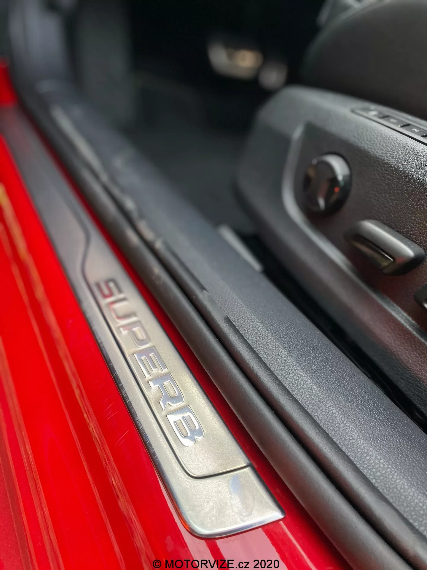 A 2019-es Skoda Superb utasoldali ajtóküszöbének közeli képe. A ferde szögből készült kép az 'SUPERB' feliratot tárja elénk a kefés rozsdamentes acél küszöbön, amely a jobb esztétikai megjelenés érdekében megvilágított. A piros külső festék éles ellentétben áll a sötét belső térrel. Az ajtó pereme műanyag burkolattal és elektromos ablakvezérléssel látható, kiemelve a fejlett funkciókat. A sekély mélységélesség kiemeli a küszöböt, bokeh hatást keltve a háttérben.