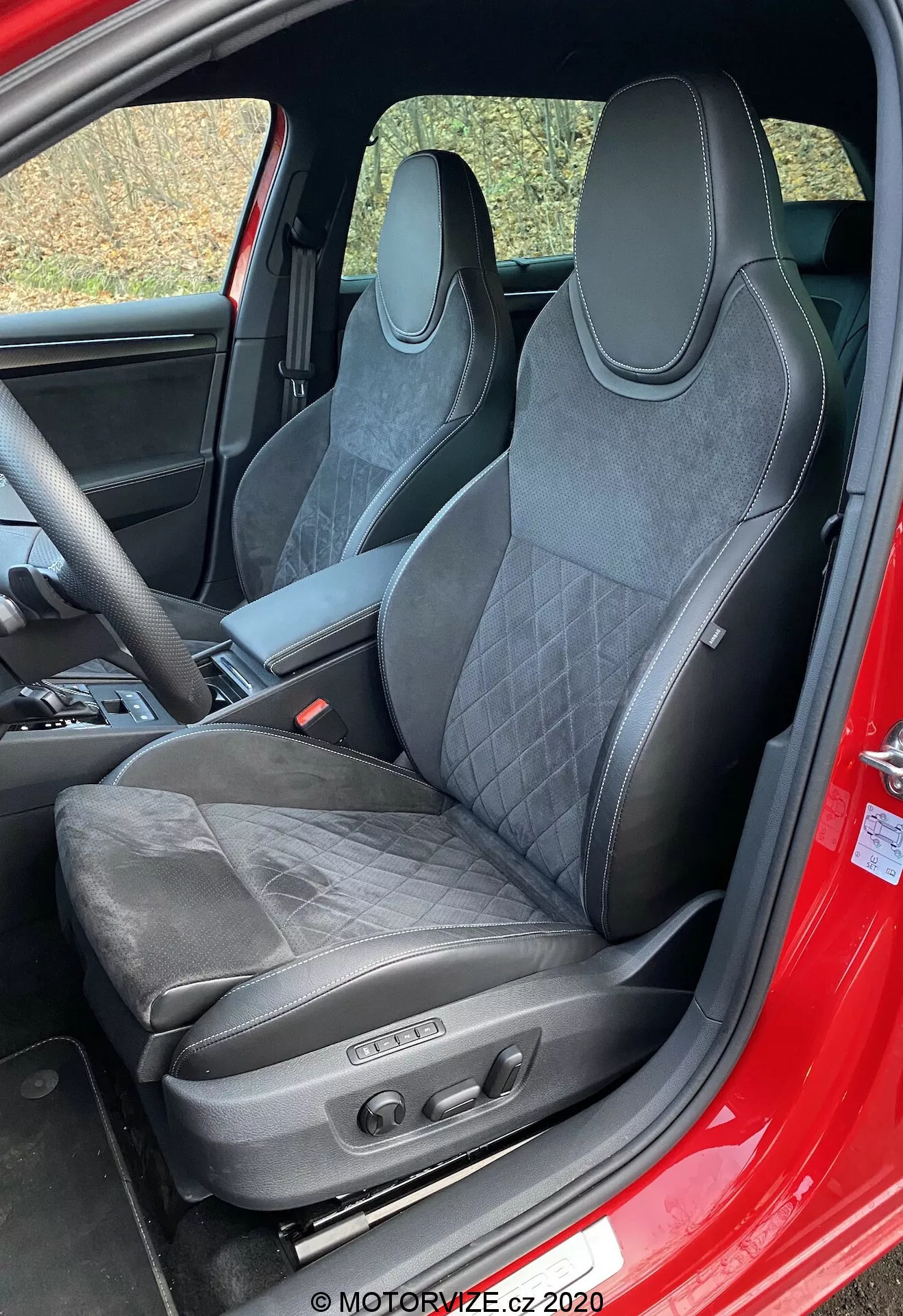 A 2019-es Škoda Superb belsejének fókuszában a vezetőülés áll. A sportos, kagylószerű ülés bőr és velúr-kárpitozással, steppelt varrással, integrált fejtámlával, kifejezett vállkontúrokkal és elektronikus állító szerkezetekkel a teljesítményorientált kialakítást, luxust és fejlett kényelmi funkciókat mutatja. A látható piros ajtókeret arra utal, hogy a külső piros lehet.