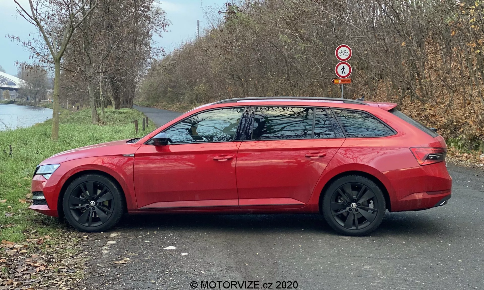 A modern 2019-es Skoda Superb kombi (Superb-Limousine) oldalsó profilképe piros színben, amelyben látható a meghosszabbított tetővonal, fekete alufelnik és a karosszéria színében lévő, beépített irányjelzőkkel ellátott oldalsó tükrök. Az autó egy aszfaltos ösvényen parkol füves terület mellett, háttérben fákkal és vízfelülettel, természetes nappali fényben.