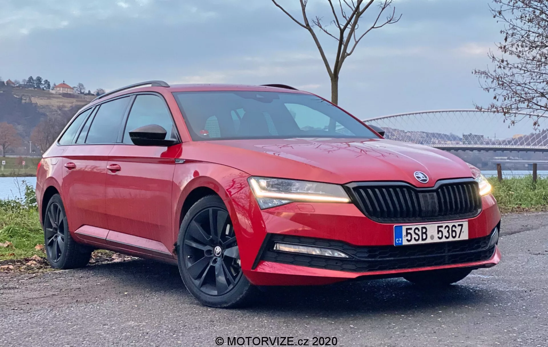 Alacsony szögből elölről negyedik nézet egy piros 2019-es Škoda Superb járműről, amely szabadtéren parkol, kiemelve a dizájnvonalakat, hűtőrácsot, kerekeket, az oldalsó profilokat és a sportos sötét színű könnyűfém felniket a festői háttérrel, vízzel és fákkal.