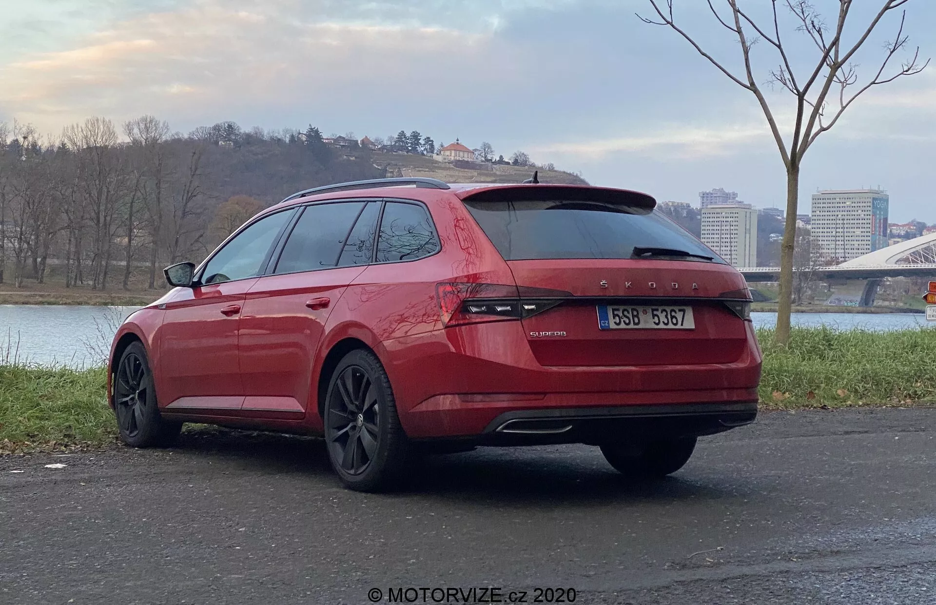 Dőlt hátsó nézet egy piros Skoda Superb 2019 kombiról, amely szabadtéren parkol, a jármű hátuljára és utasoldalára fókuszálva. Folyamatos hátsó lámpákkal a csomagtérajtó teljes hosszában, kettős kipufogócsövekkel, sötétített üvegekkel, sötét kivitelű alufelnikel, aszfaltos felületen elhelyezve, háttérben vízfelülettel és híddal.