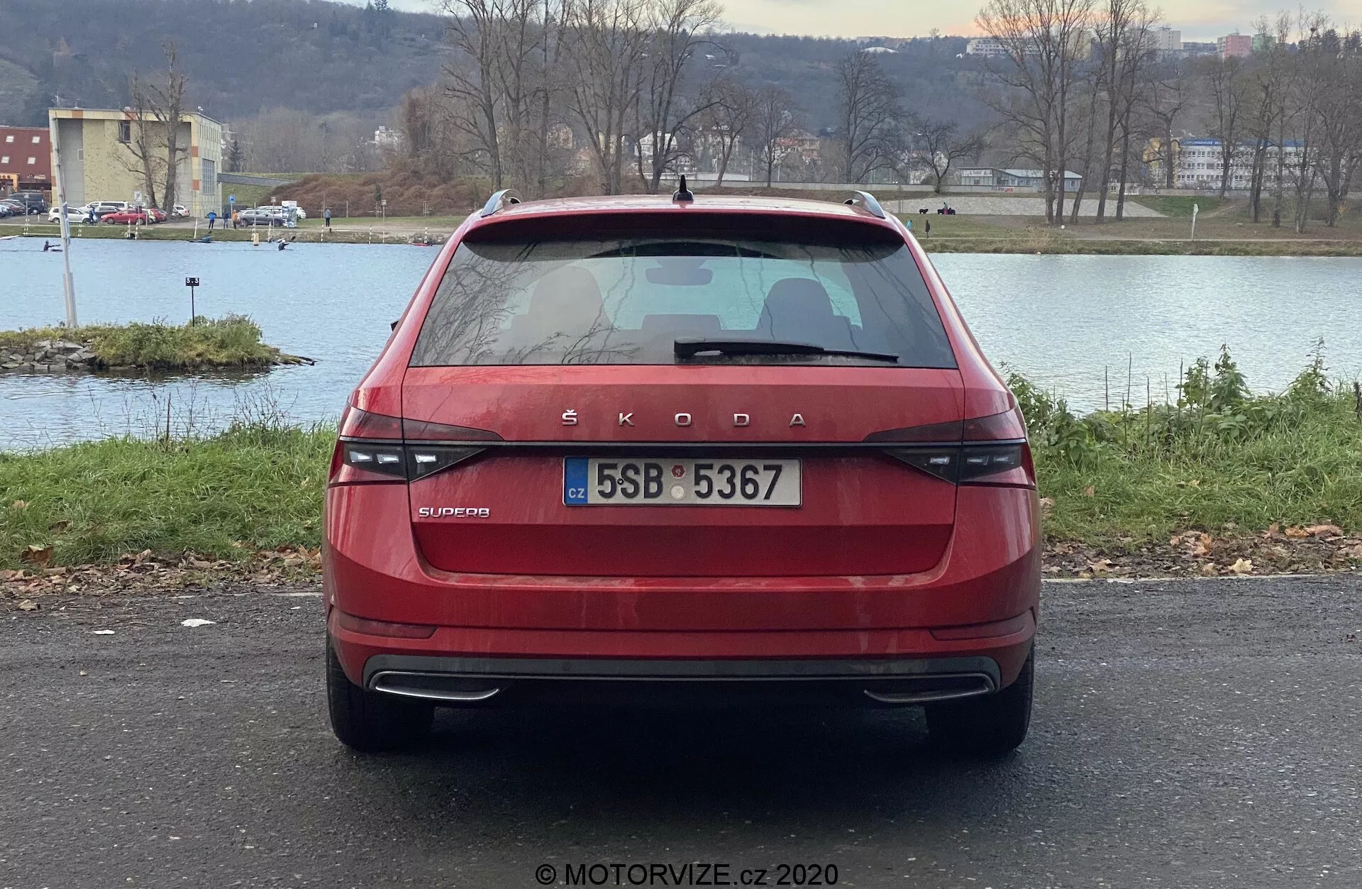 Egy piros Škoda Superb 2019 limuzin hátsó nézete, amely a jellegzetes Škoda márkajelzést és az OCTAVIA modellfeliratot mutatja a csomagtartón. A kép kiemeli a modern, vízszintes hátsó lámpákat, a központi helyzetű rendszámot, a hátsó ablaktörlőt, a színezett hátsó ablakot fagymentesítő vonalakkal, a magasan elhelyezett féklámpát, a lökhárító parkolószenzorait és a fényvisszaverőket. Az autó egy kültéri parkolóban áll, háttérben vízzel, fákkal és horizontot alkotó tájképpel.