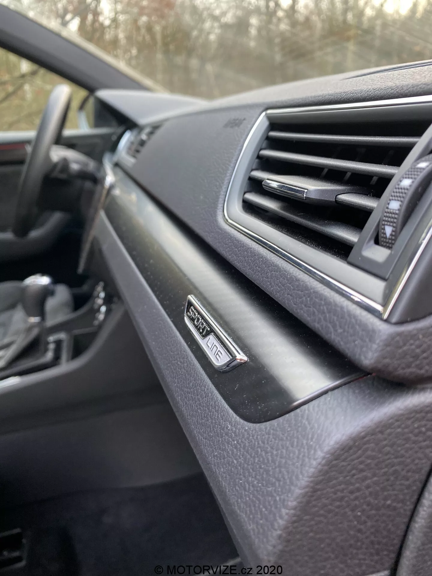 Közeli kép a 2019-es Škoda Superb belső teréről, műszerfal az utas szemszögéből. A középpontban a „Superb” jelvény a szellőzőnyílás mellett, modern dizájn puha tapintású felülettel. Elmosódott kormány a háttérben, sötét színvilág.