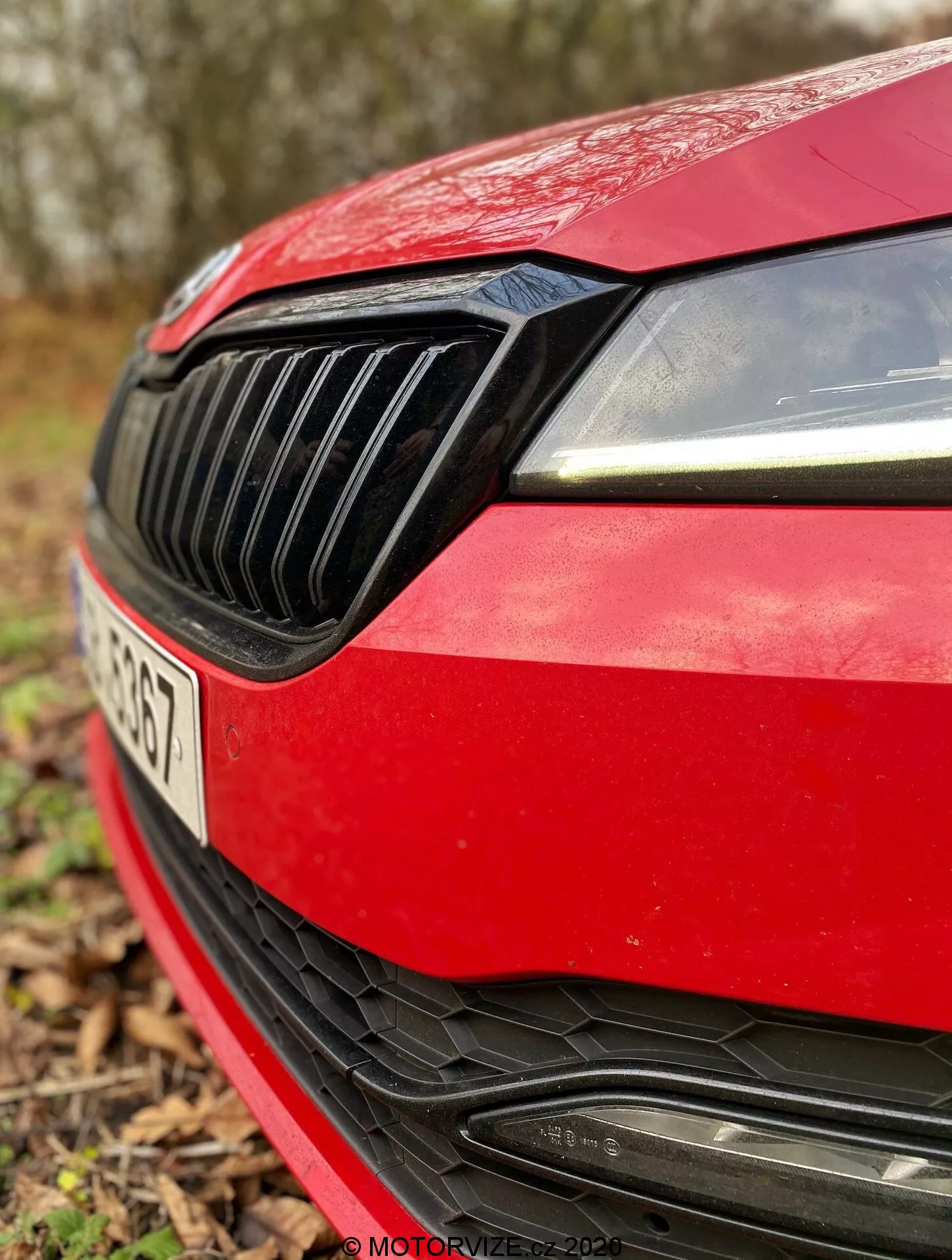 Közelkép egy piros Skoda Superb Limousine 2019-es modell bal első negyedéről. A kép a modern dizájnú jobb oldali fényszórót LED nappali világítással, a függőleges lécezéssel ellátott fekete hűtőrácsot, a karcsú piros motorháztető peremét, a sportos első lökhárítót nagy légbeömlővel és vízszintes lécekkel, valamint a gyártói embléma helyét, amely részlegesen látható. A rendszámtábla kissé látható, de a sekély mélységélesség miatt homályos, elmosódott természetes külteri háttérrel.