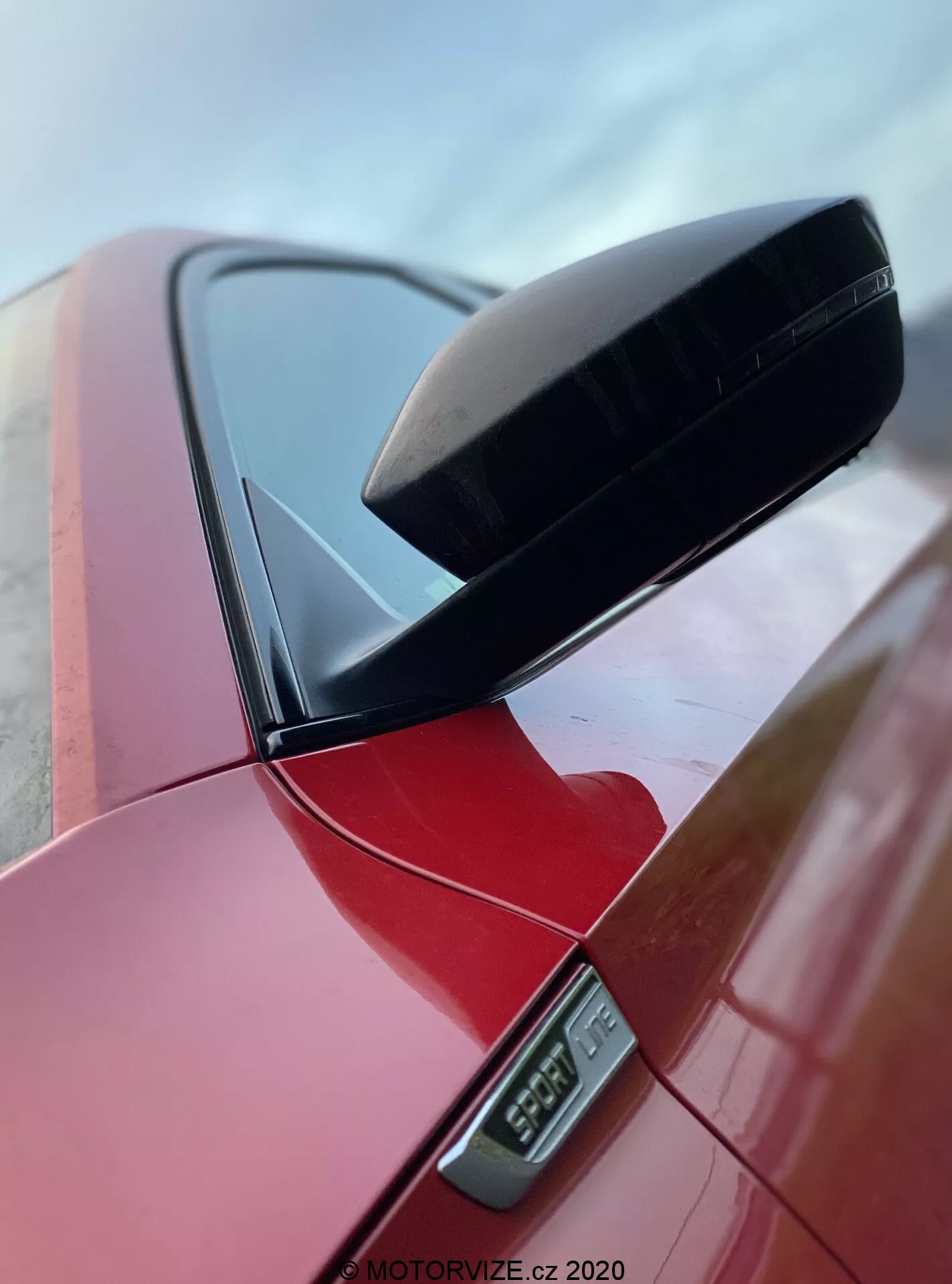 Közelkép egy Skoda Superb 2019 limuzin oldalsó visszapillantó tükréről, amely egy élénk piros színben pompázik. Ki van emelve a fekete, aerodinamikus tükörtok és az alatta elhelyezkedő LED-es oldalsó irányjelző. A kocsi egyik oszlopa látszik még, alacsony szögből készített felvétel, amely a tiszta dizájnt és a polírozott felületet emeli ki, a MOTORVIZE.cz 2020 vízjellel ellátva.
