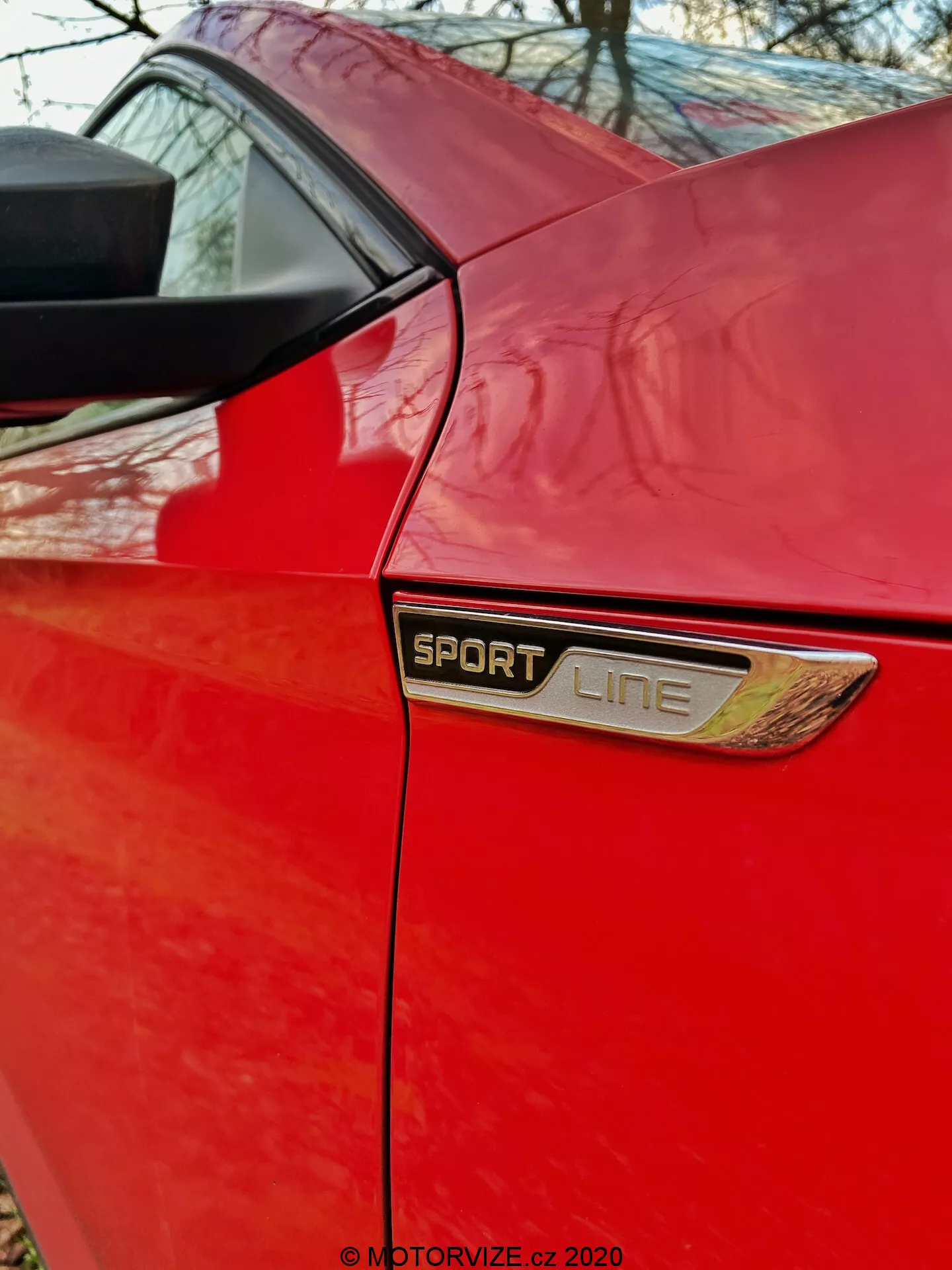 Közelkép, szögben nézett alsó negyed panel egy piros Skoda Superb 2019 limuzinon. A kép az oldalsó ajtó mögötti területre összpontosít, középen jól látható 'SPORT LINE' emblémával, krómozott vagy fémes kerettel és kiegészítő díszítő szellőzővel alatta. Az embléma fölött látható a hátsó oldalsó ablak alsó széle és a tetőszerkezetet támogató hátsó oszlop. A fotó kiemeli a jellegzetes kivitel szinteket, esztétikai elemeket és a sportos kivitel festékmennyiségét