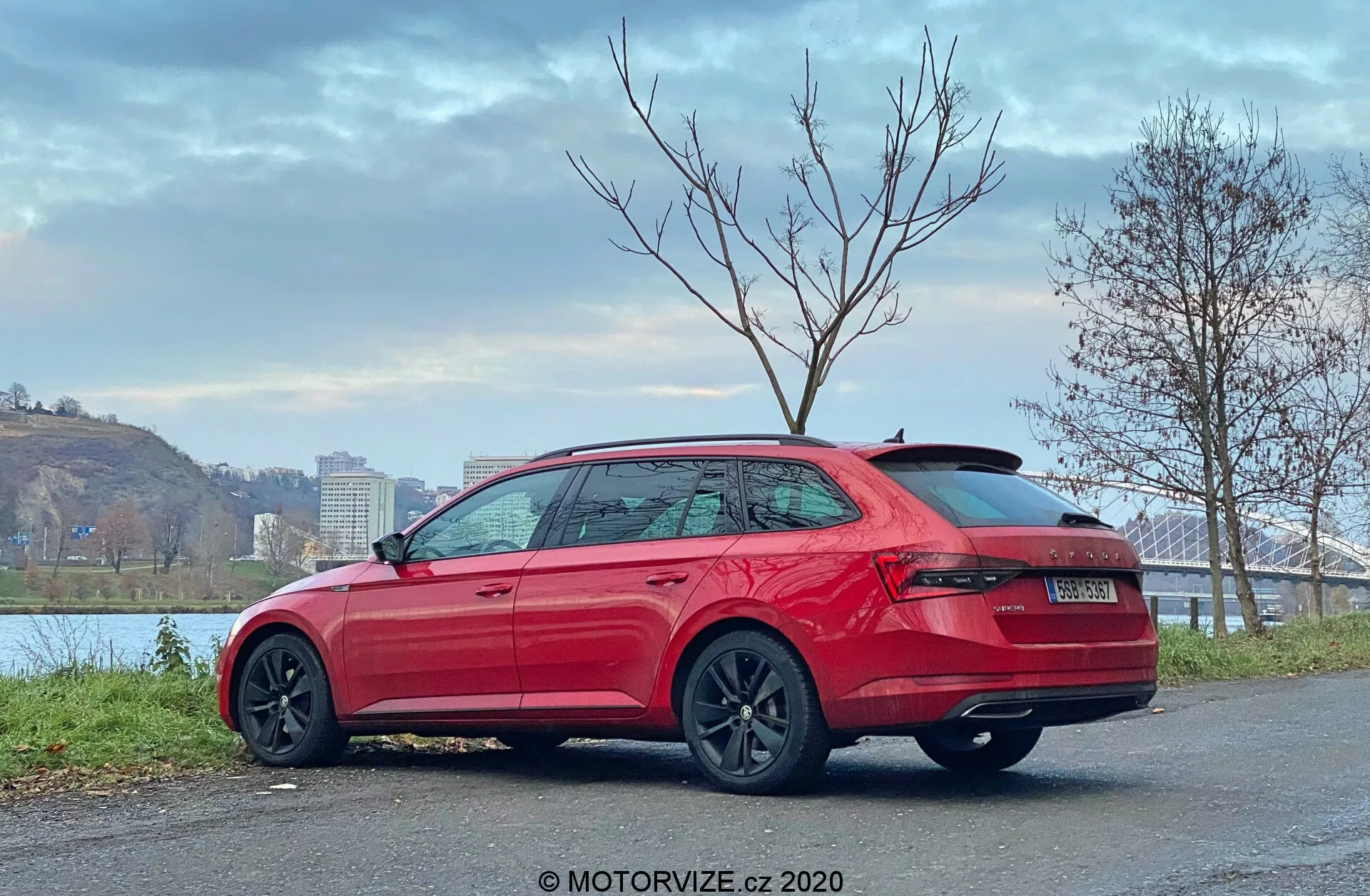 Egy 2019-es Skoda Superb kombi képe, hátsó háromnegyedes nézet, amely magasan elhelyezett hátsó lámpákat, masszív lökhárítót, látható kipufogócsövek nélküli kialakítást, tetőspoiler, elegáns oldalsó kontúrvonalakat, karosszéria színű ajtókilincseket, oldal alsó szoknyák védelmét, krómozott ablakdíszítést, sötét sokküllős kerekeket mutat, kövezett felületen parkolva a folyópart közelében híd és fák társaságában, borult égbolt, MOTORVIZE.cz 2020 vízjel.