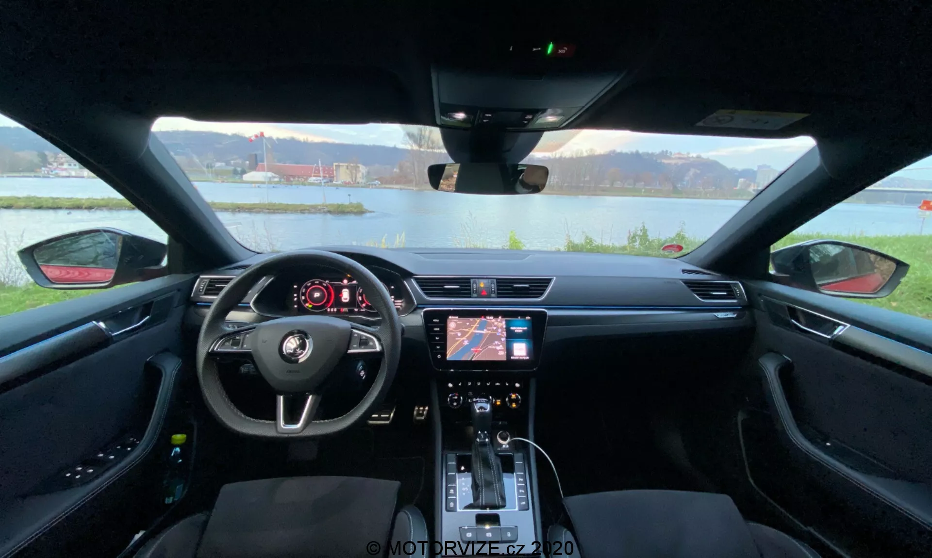 A Skoda Superb 2019 belső terének nézete a hátsó ülésről előrefelé a műszerfal felé, amely az autó belsejének teljes szélességét lefedi. Az előtérben jól látható a központi kormánykerék beépített vezérlőkkel és a Skoda logóval. A kormánykerék mögött digitális kijelző van, ami a jármű alapvető információit mutatja. A középkonzolon egy infotainment rendszer található navigációval és média beállításokkal, valamint alattuk klímavezérlők és funkciógombok. A sebességváltó kar, a pohártartók és a kontrasztos varrással ellátott részletes ülések a modern esztétikát és a vezető kényelmére és komfortjára helyezett hangsúlyt emelik ki.