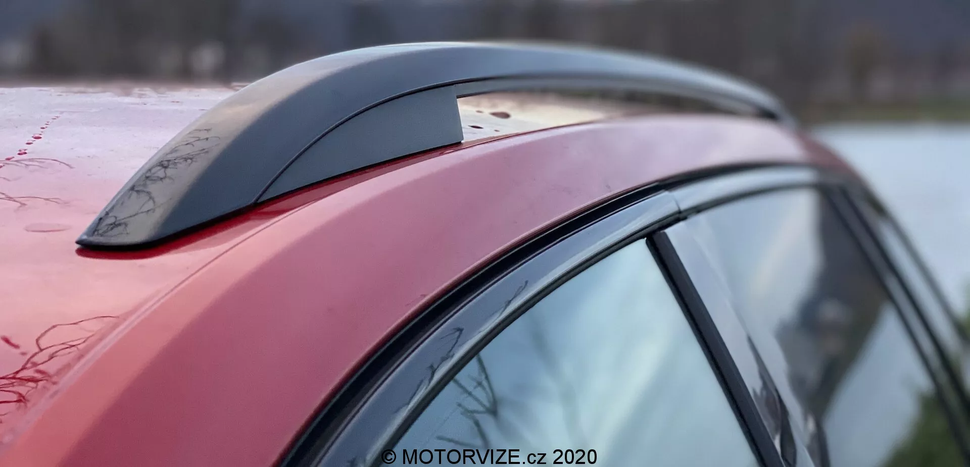 Közelről oldalnézet egy Škoda Superb 2019 jármű szekciójáról, a mélyvörös tetővonalra, az oldalsó oszlopra és a cápauszony-antenna tartozékra összpontosítva, látható vízcseppekkel, amelyek viaszos vagy hidrofób felületet jeleznek; az autó aerodinamikai kialakítása a tető ívéből és lejtéséből kivehető