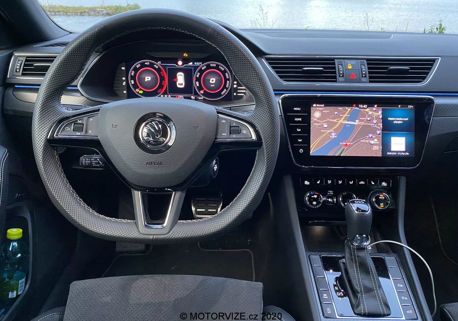 Vezetői perspektívából a Skoda Superb 2019 belseje, középen elhelyezett kormánykerékkel, mely ujjpihentetőkkel, többfunkciós gombokkal és a gyártó logójával van ellátva. A kormány mögött digitális műszerfal található, sebességmérő és fordulatszám-mérő műszerekkel, valamint további digitális kijelzőkkel, amelyek üzemeltetési részleteket mutatnak. A műszerfal közepén egy infotainment érintőképernyő helyezkedik el, alatta egy klímavezérlő rendszer gombokkal és forgókapcsolókkal. Az alsó középkonzolon automata váltó karja, elektronikus kézifék gombja és vezérlő gomb található. A vezetői oldal ajtója és a visszapillantó tükör is látható, az ajtó kezelőszervei észrevehetők. Az utastér különböző anyagok, krómozott díszítések, bőr borítású kormánykerék, valamint fényes fekete műanyag kombinációját használja, modern esztétikát biztosítva.