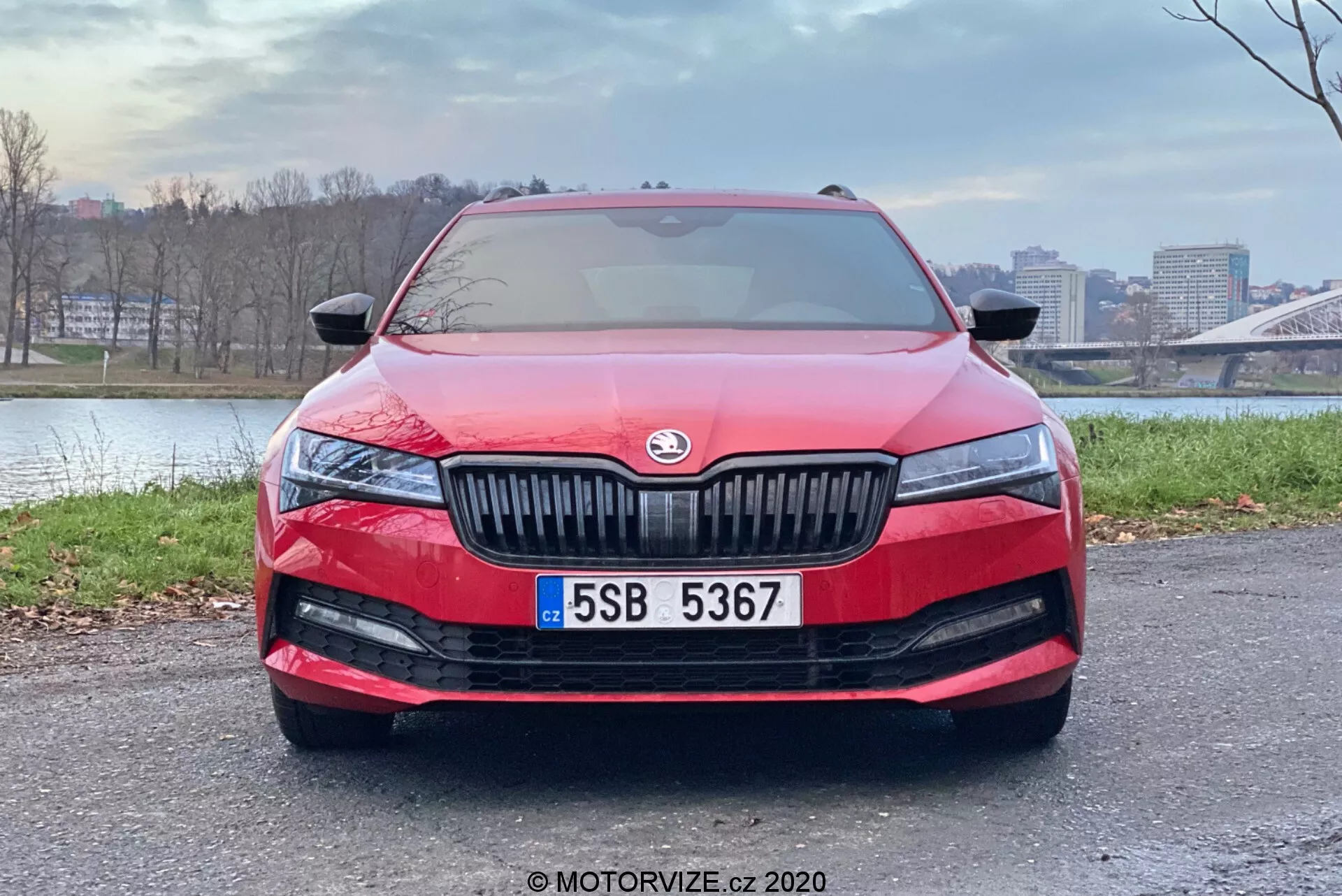 Modern piros Skoda Superb Limousine 2019 elölnézete, amely bemutatja sima aerodinamikus kontúrokat, a hangsúlyos függőleges rácsot a központilag elhelyezett gyártói emblémával, a karcsú és meghosszabbított LED-es fényszórókat, az alsó lökhárító széles légbeömlő rácsát ködlámpákkal, a központilag elhelyezett rendszámtáblát és esetleg egy érzékelőt vagy kamerát a fejlett vezetőtámogató rendszerekhez. A szélvédő tükrözi az eget, jelezve az esőérzékelős ablaktörlőket vagy fényszenzort az automatikus fényszóró aktiváláshoz. Az autó egy burkolt területen parkol városi háttérrel, kiemelve a dizájnját és funkcióit.