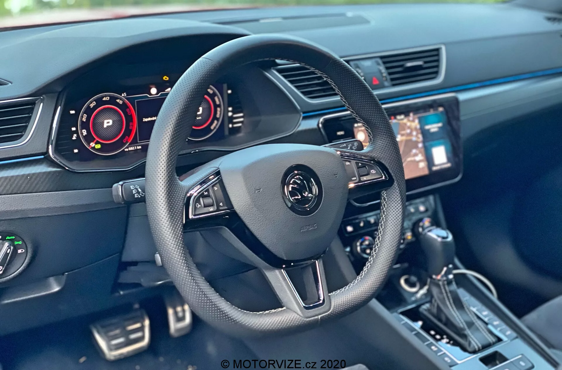 Skoda Superb 2019 belső tere a vezető üléséből nézve, modern, háromküllős kormánykerékkel, multifunkciós vezérlőkkel és központi emblémával. A látható alkatrészek közé tartozik a digitális műszerfal, központi infotainment rendszer érintőképernyővel, konzol, sebességváltó kar, gombok és klímavezérlő rendszer. Az autó belső technológiája olyan elemeket mutat, mint a digitális műszerfalak és a kormánykerék vezérlés.