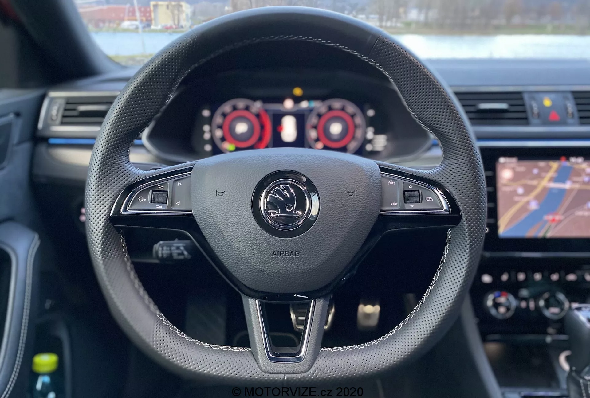 A 2019-es Skoda Superb Limousine belső nézete a vezető szemszögéből. A vezetőülés egy fekete bőrrel bevont, varrással díszített kormánykereket tár fel, amely különféle vezérlőket tartalmaz, beleértve a hangerő-szabályozást és valószínűleg a jármű beállításait. Az kép közepén látható a gyártó emblémája az 'AIRBAG' felirattal, ami a vezető oldali első légzsák jelenlétét hangsúlyozza. A kormánykerék mögött két váltófül is látható, ami manuális sebességváltást jelez. A műszerfal, rajta a fordulatszámmérővel, sebességmérővel és digitális kijelzővel, részben látható, míg az infotainment rendszer navigációs térképet jelenít meg. A sekély mélységélesség a kormánykerékre és az emblémára fókuszál, miközben a műszerfal és az érintőképernyő enyhén elmosódott.