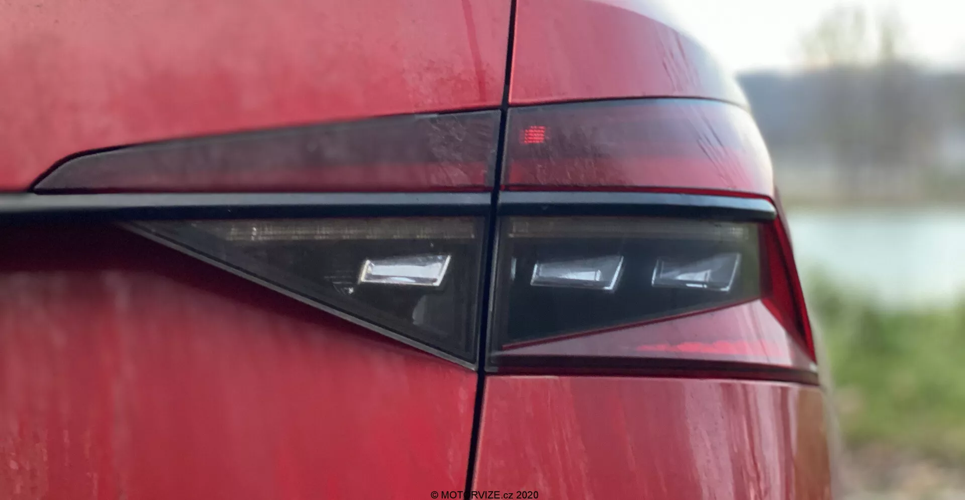 A Skoda Superb 2019 hátsó részének közeli képe, amely az oldalsó szögből vett hátsó lámpa egységére összpontosít, és bemutatja a modern, elegáns kialakítást vízszintes LED fényekkel, sötétvörös lencsébe burkolva, tükrözve a kortárs sportos vagy luxusautók esztétikáját. A fényes karosszéria és a fekete vízszintes díszléc növeli a vizuális vonzerőt a homályos háttér előtt.