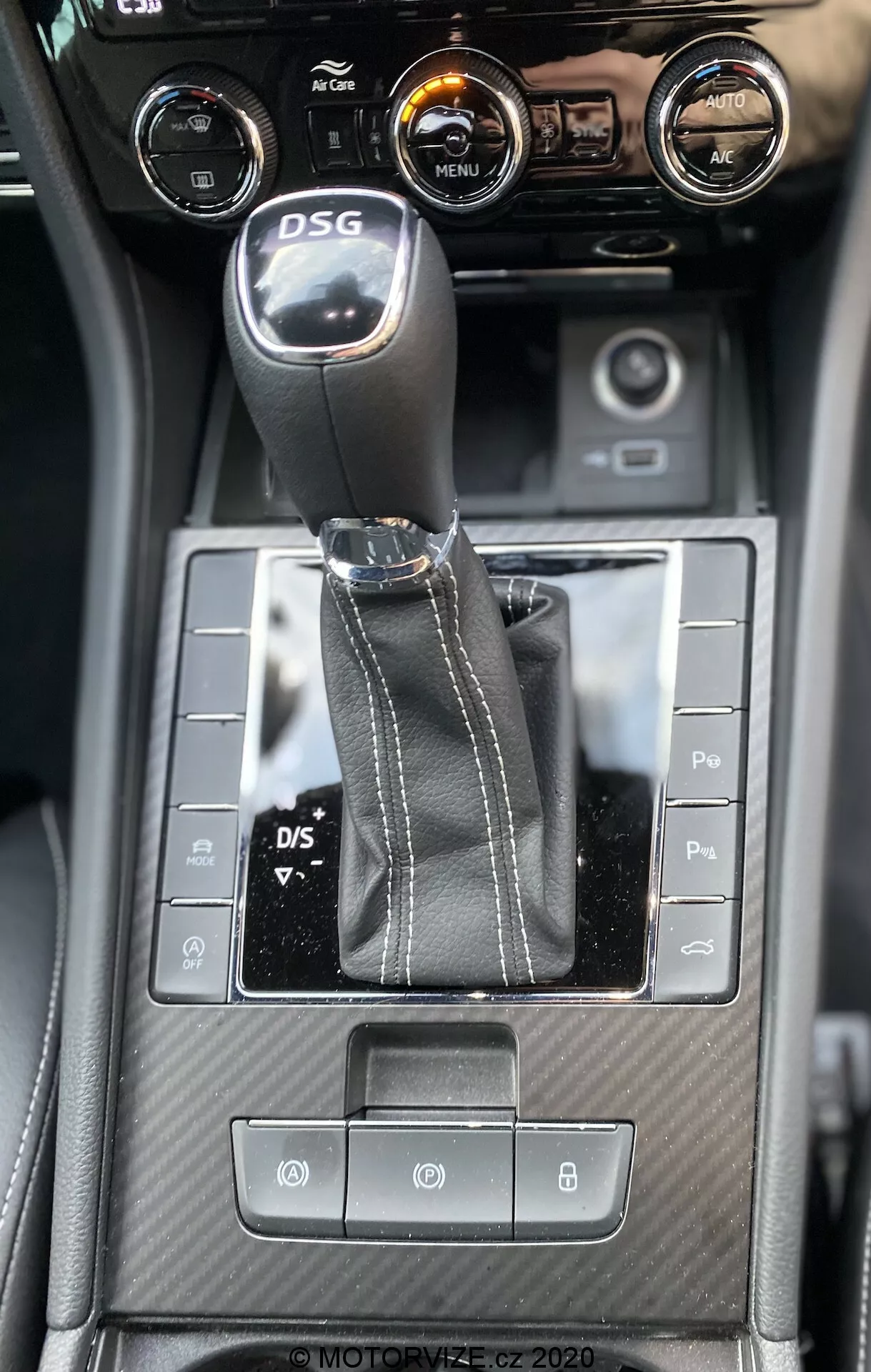 A Skoda Superb 2019 belső középkonzoljának belső nézete lefelé néző szögből, kiemelve a DSG sebességváltó kart, szimmetrikus vezérlőgombokat, karbon-szálas hatású textúrázott részleteket és a klímaberendezés vezérlőit központi érintőkijelzővel, ami járműinformációkat jelenít meg.