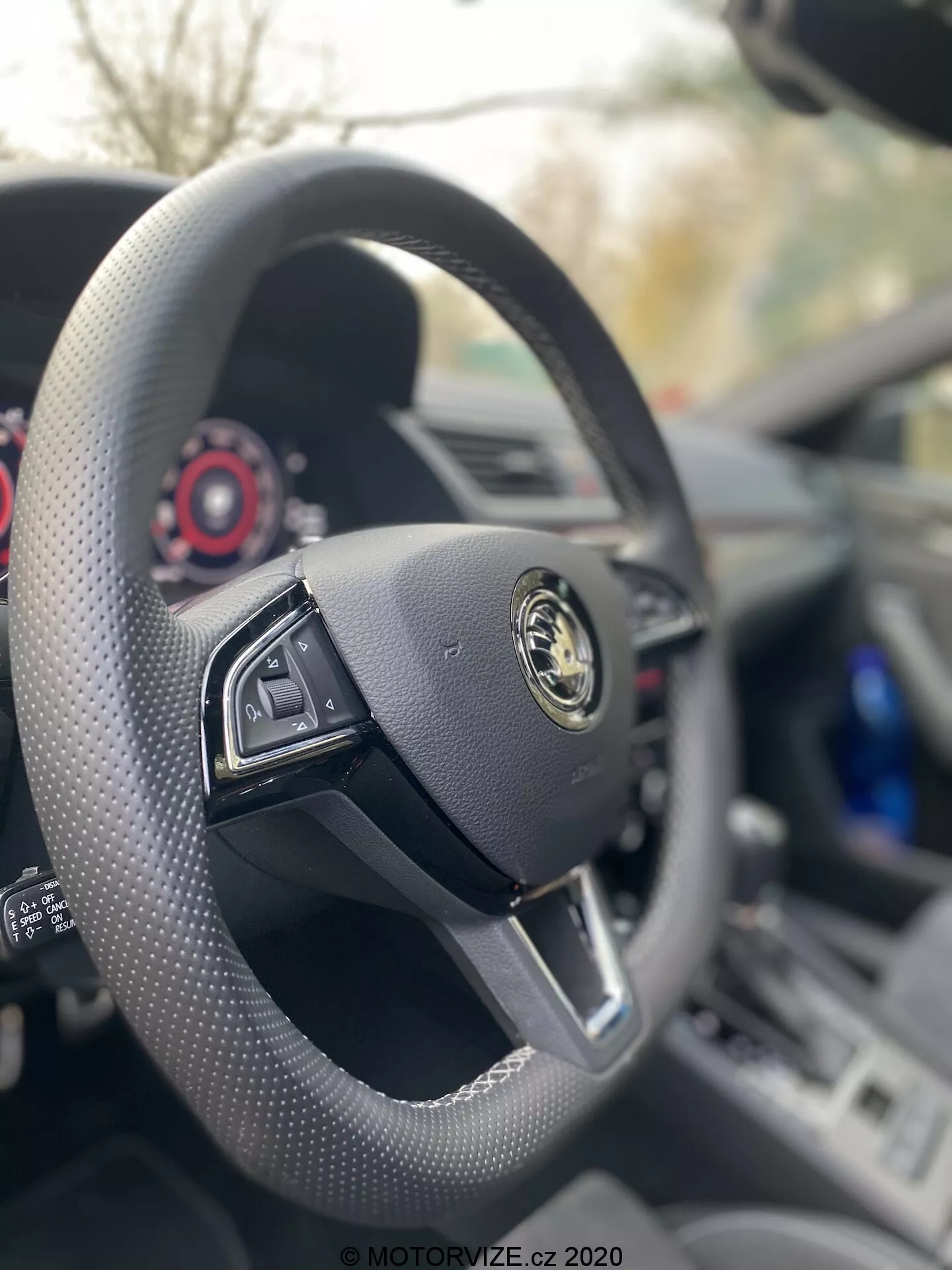 A 2019-es Skoda Superb Limousine belső nézete a vezetőülésből, fókuszban a multifunkcionális gombokkal ellátott fekete bőrborítású, lapos aljú kormánykerékkel. A gyártó logója, arany emblémával a fekete háttérrel, jól láthatóan kiemelkedik a közepén. A műszerfal és a műszercsoport látható a kormány mögött, kiemelve az autó minimalista, mégis luxus kialakítását, piros részletekkel az aktuális számlapokon.