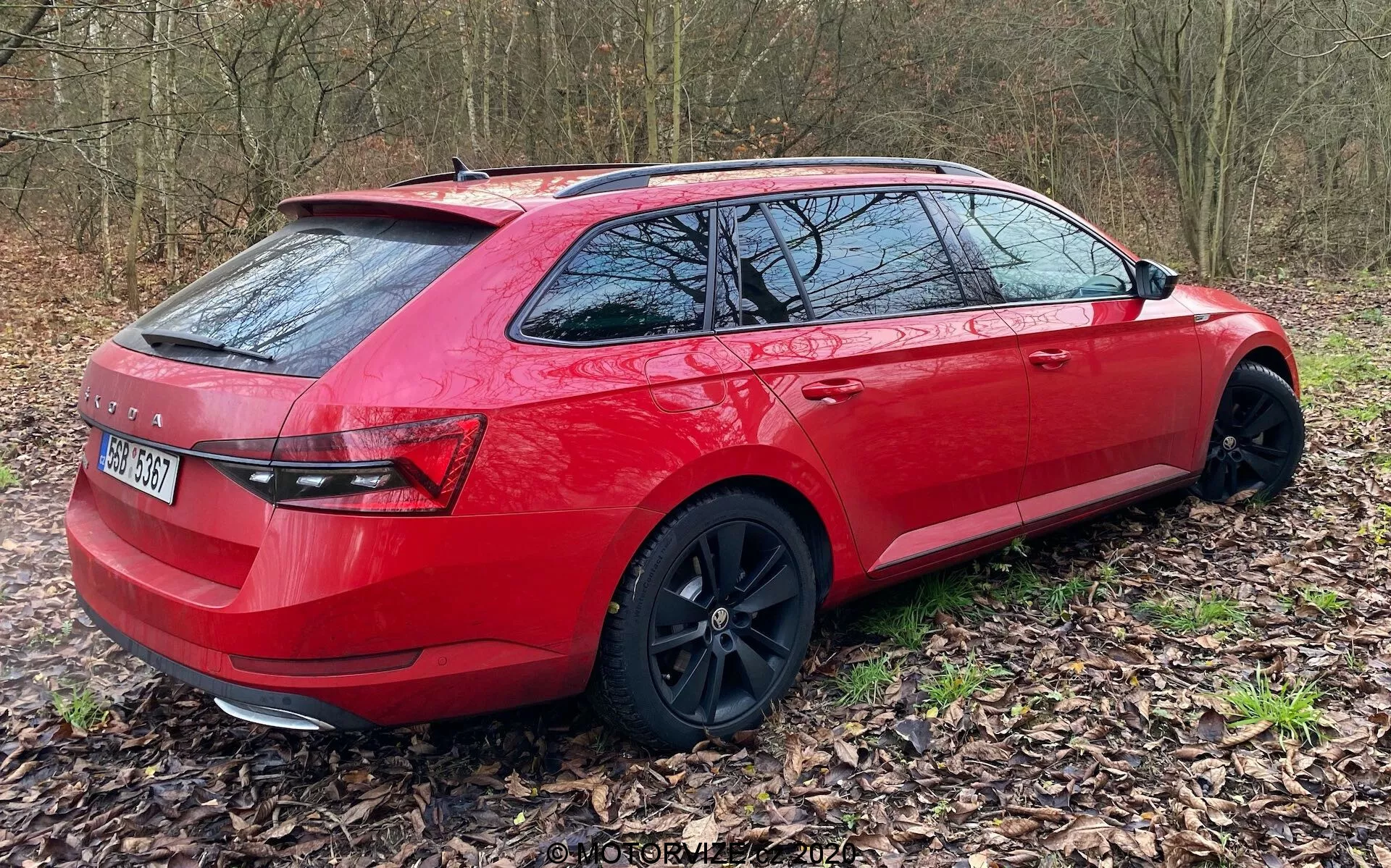Egy piros kombi háromnegyedes hátsó nézetből, amely ágak és levélszőnyeggel borított természetes terepen van parkolva. Az autó jobb oldala a hátulsó lökhárítótól az első ajtókig látható, bemutatva a modern, prémium dizájnt. A fekete könnyűfém kerekek kissé jobbra fordulnak, sportos és elegáns megjelenést kölcsönözve a járműnek. A jellemzők közé tartoznak a modern hátsó lámpák, alacsony profilú gumik és a tetősín rendszer. A sötétített hátsó ablakok fokozott magánéletet és napvédelmet sugallnak.