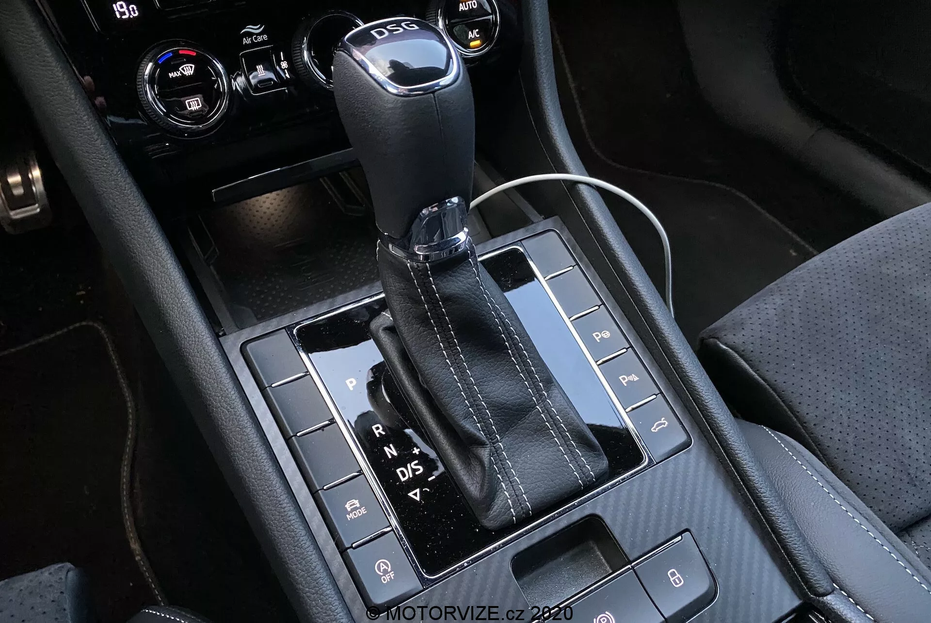 Közelkép a Skoda Superb 2019 automata sebességváltó karjáról és a környező középkonzolról, bőrrel bevont, fém tetejű váltókarral, látható 'P, R, N, D/S' betűkkel, amelyek jelzik a különböző sebességeket. A fényes fekete panel számos gombot tartalmaz, mint például parkolásérzékelők, Auto Start/Stop rendszer kikapcsolása és kipörgésgátló. A konzolon anyagok keveréke látható részletes varrással, beleértve egy 12 voltos csatlakozót és valószínűleg klímaberendezési vagy infotainment vezérlőket a kivágott jobb felső sarokban.