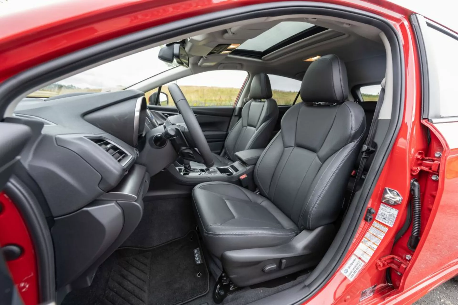 2018-as Subaru Impreza belső nézete külső perspektívából, amely megmutatja a vezető ülését, a műszerfalat, a kormánykereket vezérlőgombokkal, az ergonomikus fekete bőr első üléseket és az ajtó belső vezérlőit a vezető ajtaján keresztül nyitva