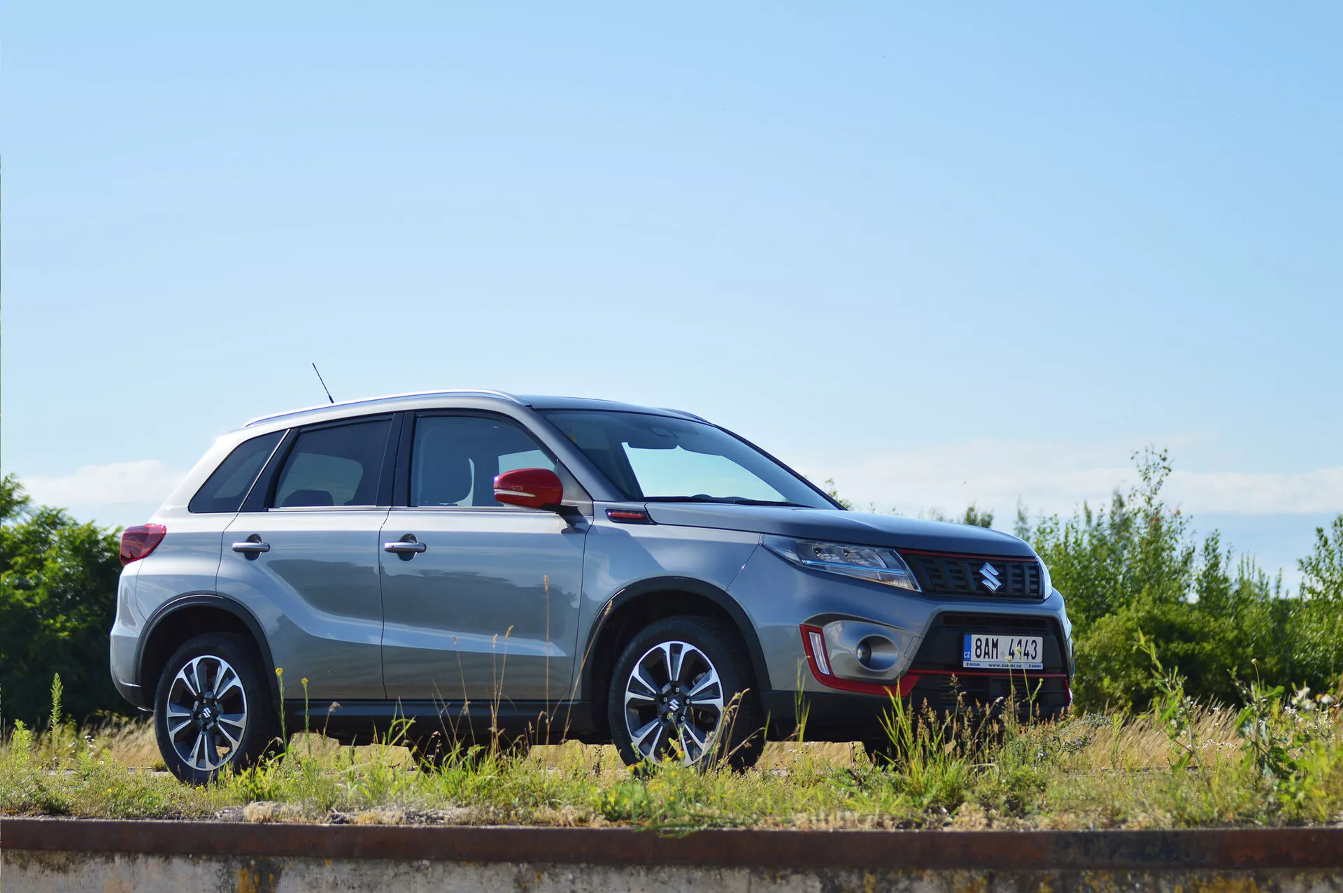 Oldalsó profil nézet egy ezüstszínű 2018-as Suzuki Vitara kompakt SUV-ról, amely párhuzamosan parkol egy vasúti sínnel egy napos napon. Egyértelműen láthatók a dizájnvonalak, a többküllős alufelnik, a metálfényezés, a zárt ajtók és ablakok, a fényszóró alakja, az integrált indexlámpák lehetőségével ellátott tükör, valamint a felszereltségi szint jelvénye, amelyek a jármű dizájnját és technológiai jellemzőit mutatják be.