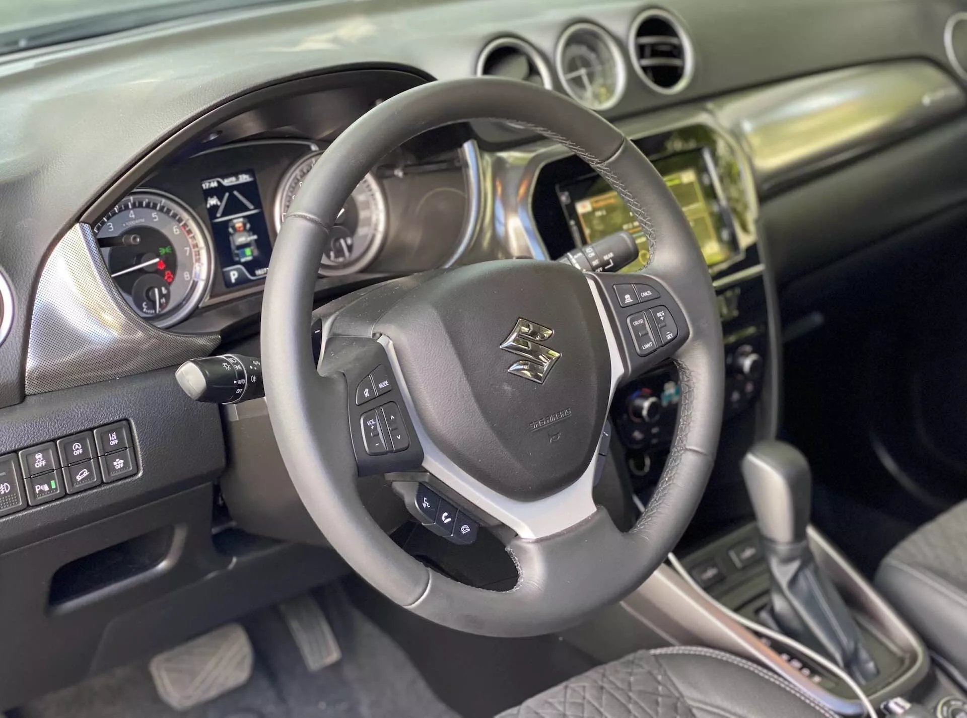 A 2018-as Suzuki Vitara belső tere, a vezető perspektívájának rögzítése. Közeli kép egy bőrrel bevont, többfunkciós kormánykerékről emblémával a középpontban, beépített hangvezérlési, hangvezérlési és sebességtartási rendszerrel és fémes ékezetekkel. Mögötte egy műszerfal található analóg sebességmérővel és fordulatszámmérővel, és egy digitális kijelzővel a jármű információihoz, ami azt sugallja, hogy az autó kikapcsolt állapotban vagy tartozék módban van. A középkonzol egy fém- vagy krómfinomított halmot mutat érintőképernyős infotainment rendszerrel, gombokkal és szabályzókkal a klímaberendezés és a jármű vezérléséhez. Az automata váltó bőrborítású ékei különböző vezetési módokra utalnak. A belső tér bőr és csiszolt fém kombinációját kínálja, tükrözve a modern dizájnt. Feltételezhető haladó funkciók közé tartozik a Bluetooth, a navigáció, a parkolóasszisztensek és a vezetőtámogató rendszerek.