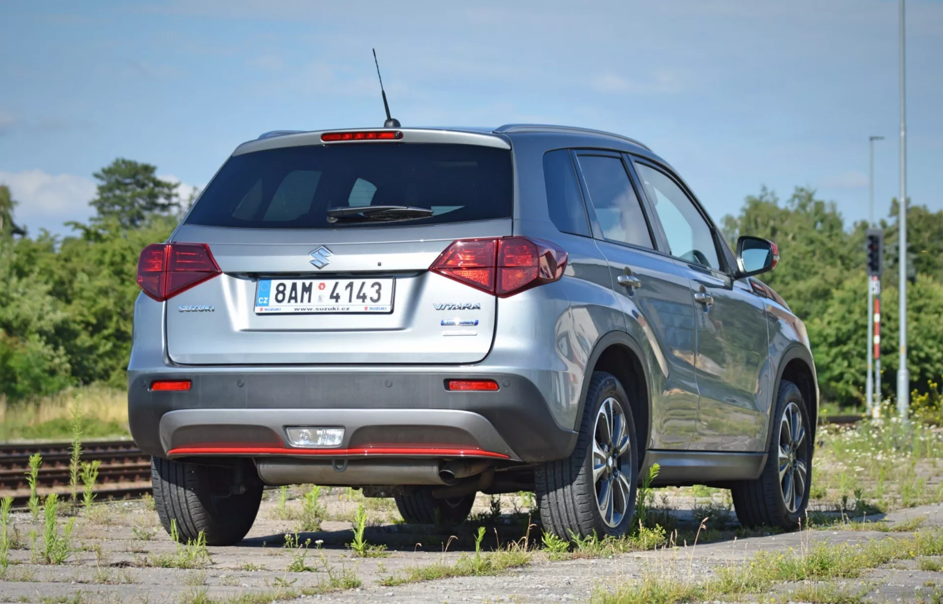 Suzuki Vitara 2018 SUV hátsó nézete alacsony szögből, kiemelve a magasságát és a hasmagasságát. 45 fokos szögben elhelyezkedő kép, amely megmutatja a hátsó ajtót modell jelöléssel, hátsó szélvédőt ablaktörlővel, hagyományos halogén hátsó lámpákat, fényvisszaverős hátsó lökhárítót és látható kipufogócsövet. A jobb oldali profil tartalmazza a hátsó utasajtót, oldalsó ablakokat, tetősíneket és a szokásos könnyűfém kerekeket. Az oldalsó tükrökre nem láthatók irányjelzők. A jármű poros felületen parkol a vasúti sínek mellett, száraz, napos területen zöld növényzet és kék ég mellett.