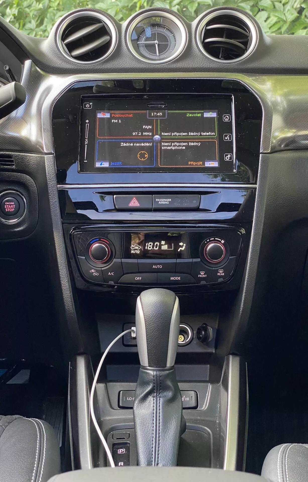 A 2018-as Suzuki Vitara belső terének részletes nézete, a középkonzolra és a műszerfalra összpontosítva az első ülések perspektívájából. A kép kiemeli az infotainment érintőképernyőt, amely egy menüt jelenít meg, a kétzónás klímaberendezés digitális kijelzőit és az automata váltó karját. A belső tér sötét színű befejezésű, matt és fényes textúrák keverékével, illusztrálva a minőségi anyagokat és a modern autóipari technológiát.