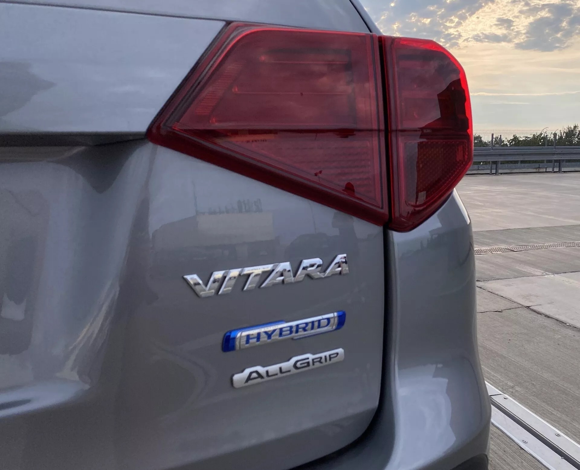 Közelkép a Suzuki Vitara 2018 bal hátsó részéről, amely bemutatja a bonyolult LED hátsó lámpa kialakítást és három jelzést, amelyek a 'VITARA' modellt, a hibrid technológiát 'HYBRID' és az összkerékhajtási rendszert 'ALLGRIP' jelzik, hajnalban vagy alkonyatkor, egy nyitott parkoló területen.