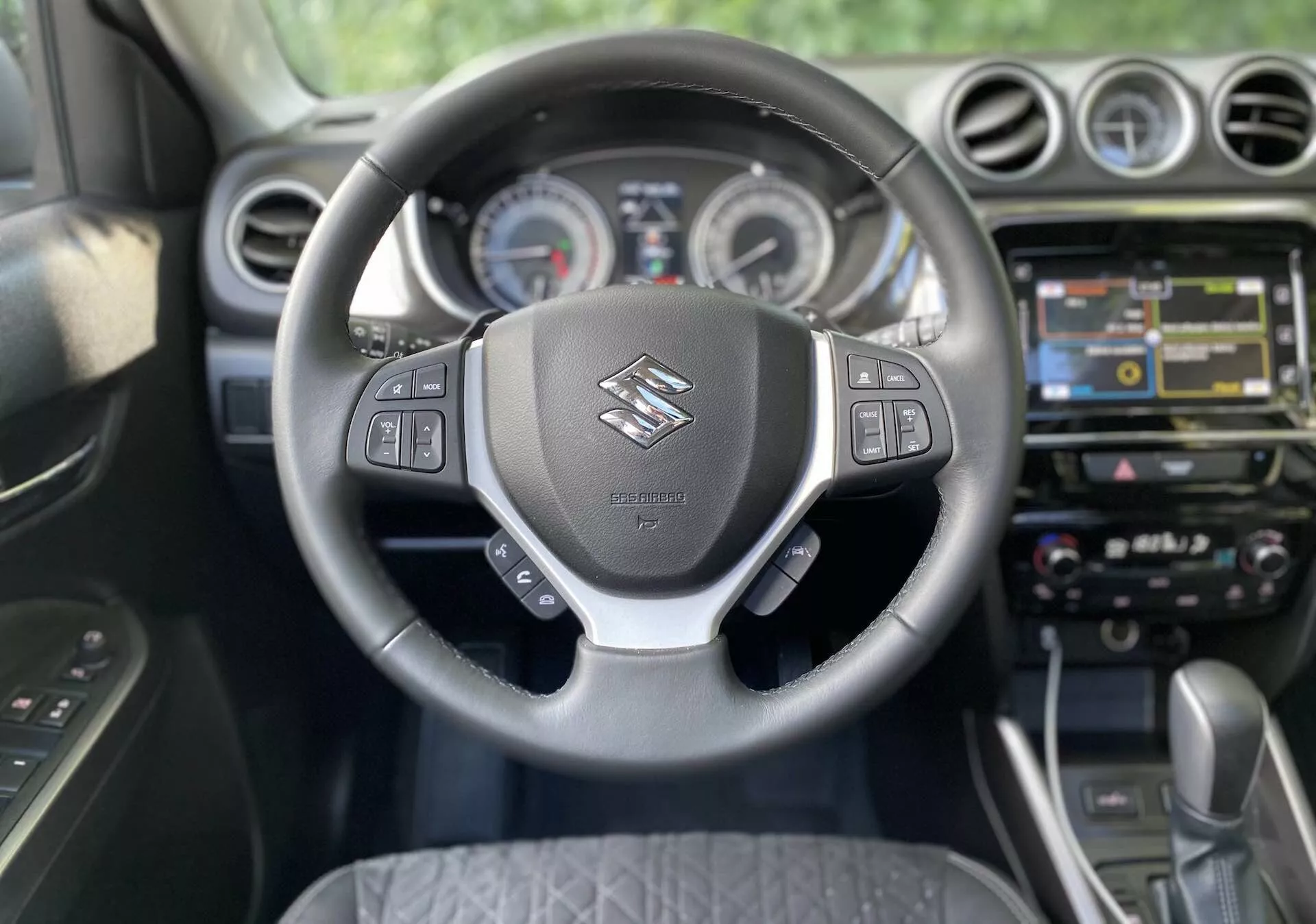 Suzuki Vitara 2018 belső nézet a vezetői ülés perspektívájából. A fókusz a kormánykeréken van, amelyen a Suzuki logó és multifunkciós vezérlők találhatók az audió, hangparancsok, sebességtartó automatika és a jármű információs kijelzőjének kezelése érdekében. A kormány mögött analóg számlapok és egy központi kijelző látható a műszerfalon. A középkonzolon érintőképernyős infotainment rendszer található navigációval, multimédiával és csatlakozási lehetőségekkel. Ezen kívül érintőgombok és tekerőgombok találhatók a kétzónás klímaberendezéshez. Az autó automata váltóval van felszerelve, ami modern és esetleg prémium kabintervet jelez.