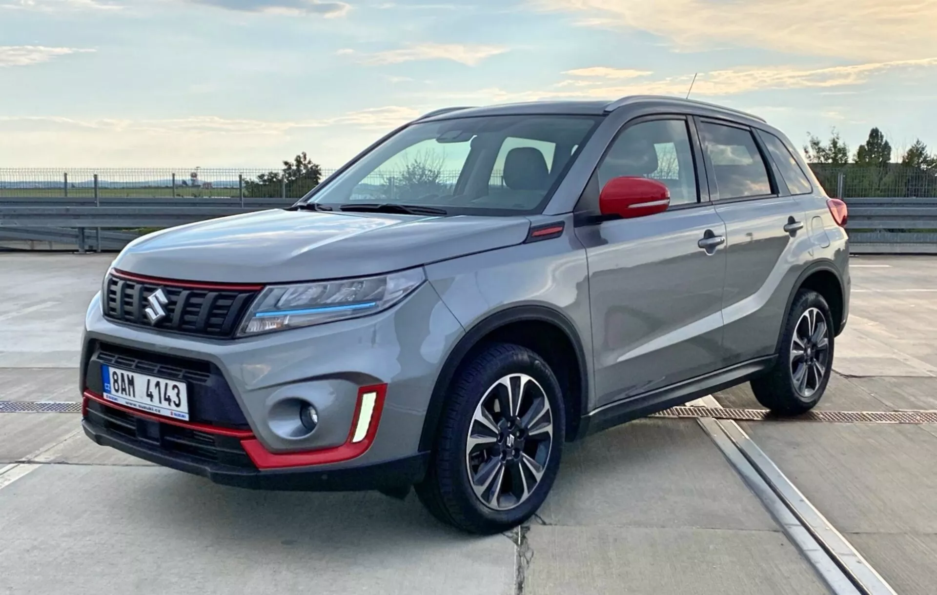 A képen egy 2018-as Suzuki Vitara kompakt SUV látható, amely egy kültéri környezetben parkol, valószínűleg egy parkoló fedélzeten, napkeltekor vagy napnyugtakor. A fotó háromnegyedes elülső szögből ábrázolja az autót, megmutatva a vezető oldalát és az elejét, kiemelve a fényszóró konfigurációját, a hűtőrács stílusát, az első lökhárítót, a kerekeket és az oldalsó karosszérialíneket. A jármű kéttónusú fényezéssel rendelkezik: az ezüstszürke test és a fekete tető, valamint az oldalsó tükrök, a lökhárítón található piros részletekkel. Elegáns LED fényszórók, egy kiemelkedő márkajelzés a mintás rácson és többküllős könnyűfém kerekek is láthatók, amelyek bemutatják a jármű modern és sportos kialakítását.