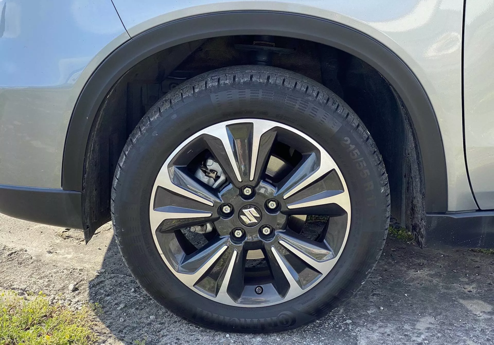 Közelkép a 2018-as Suzuki Vitara kerekéről, bemutatva egy többszörösen küllős, fémes és sötétebb festésű könnyűfém kereket. A márkázott középsapka takarja a kerékcsavarokat, és az úthasználatra készült gumiabroncs részletes futófelületi mintázatokat és oldalfali jelöléseket mutat. A háttérben a kerék feléhez tartozó sárvédős és oldalsó panel egy semleges, fényes színben látható, enyhén felfelé szögből, a talajszintről nézve.