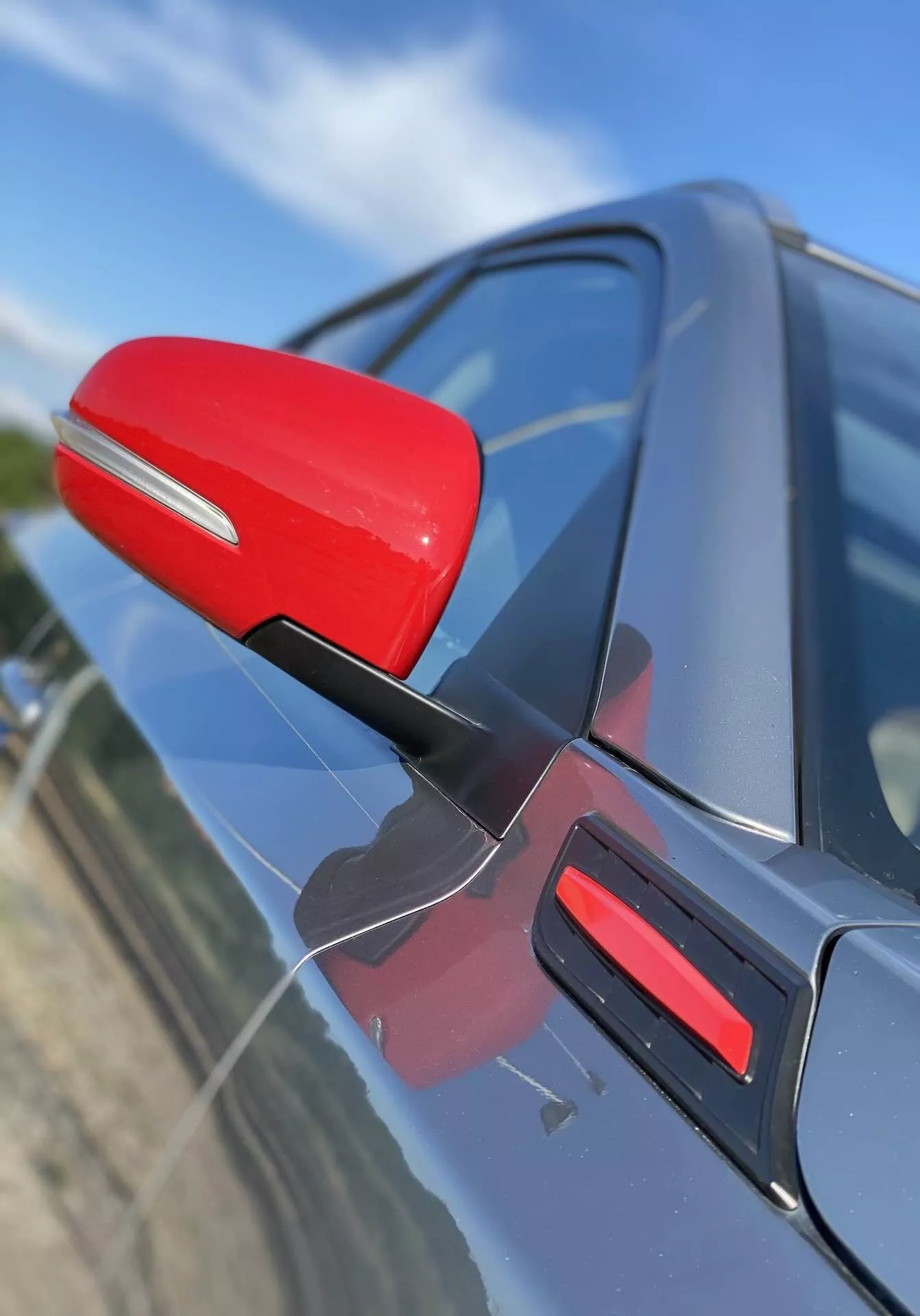 A 2018-as Suzuki Vitara sofőr oldali visszapillantó tükrének közeli képe, amely piros színnel kiemelkedik a sötét színű karosszéria ellenében. A tükör egy karcsú fekete tartóval van rögzítve, amely az aerodinamikai hatékonyságot sugallja. A kép egy kissé előre mutató, ferde szögből rögzíti a tükröt, lefelé néző perspektívával, az ajtón lévő megfelelő piros díszítéssel. A háttér elmosódott, kiemelve a tükör és az autó ajtajának design részleteit.